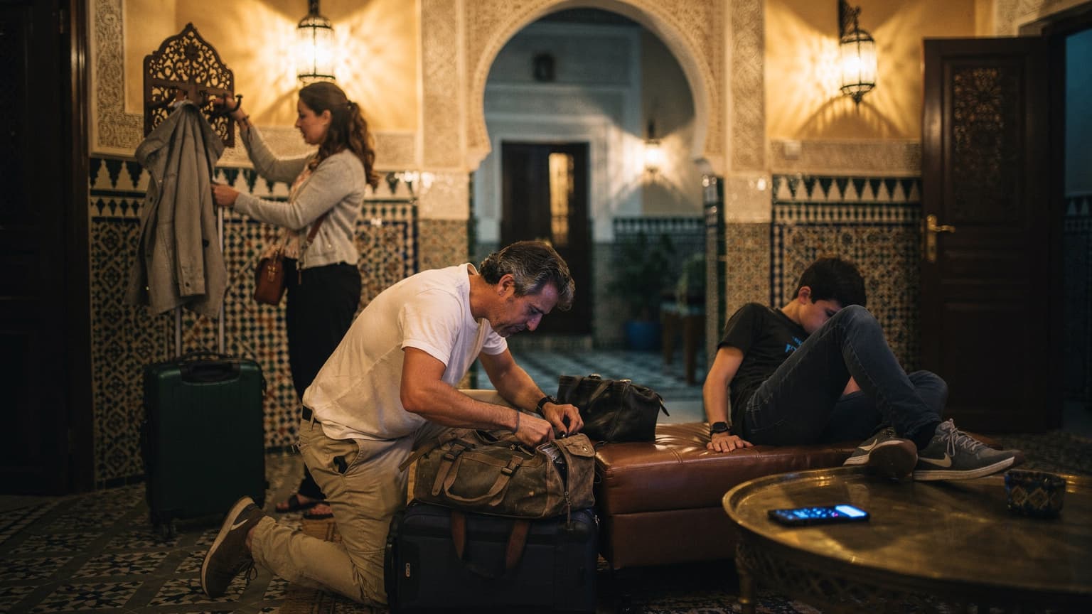 Famille en train de déballer ses bagages dans un riad à Marrakech le soir, lumière tamisée du patio, téléphone posé sur la table sans être regardé