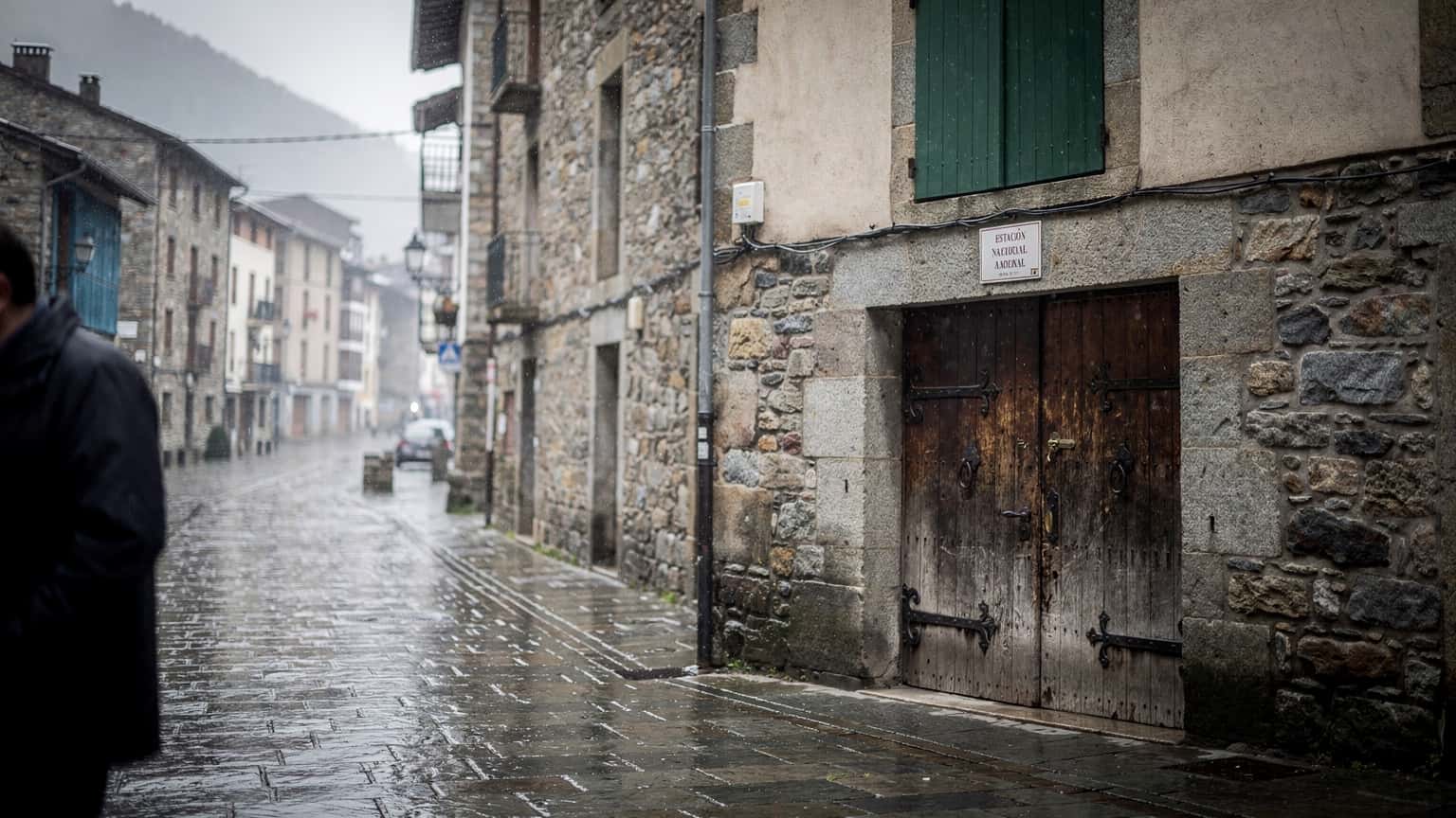 Rua molhada em Andorra la Vella com persianas de madeira e uma porta de pedra, luz cinzenta de chuva, perto da estação de comboios.