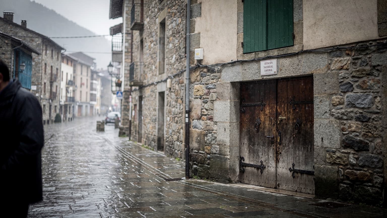 Rua molhada em Andorra la Vella com persianas de madeira e uma porta de pedra, luz cinzenta de chuva, perto da estação de comboios.