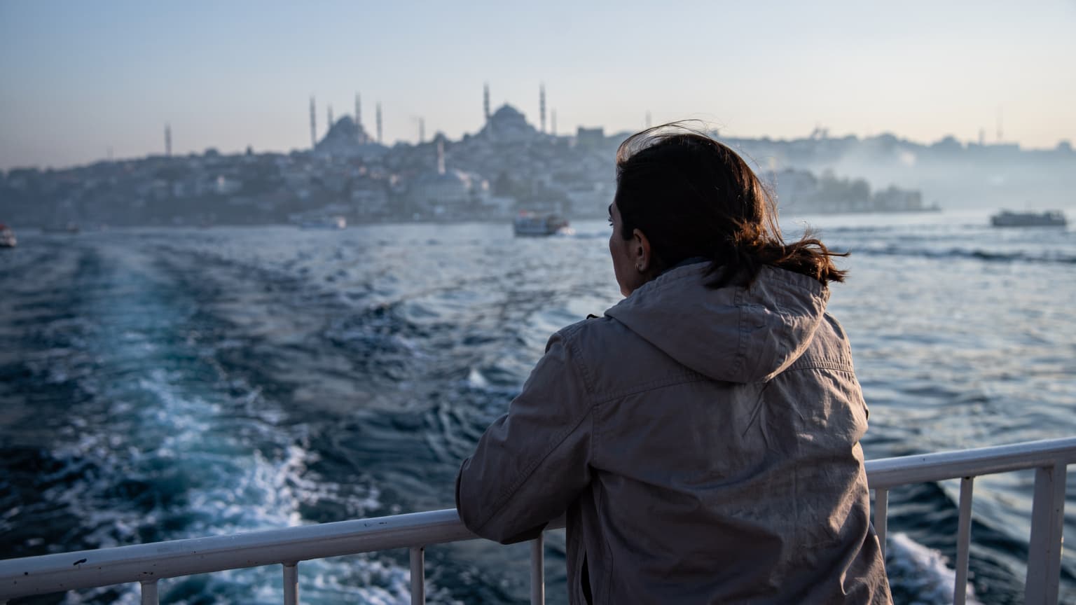 A lone figure leaning on the railing of a ferry boat, facing the sea, with the sun just beginning to rise over the distant coastline of Istanbul, casting cold blue light.