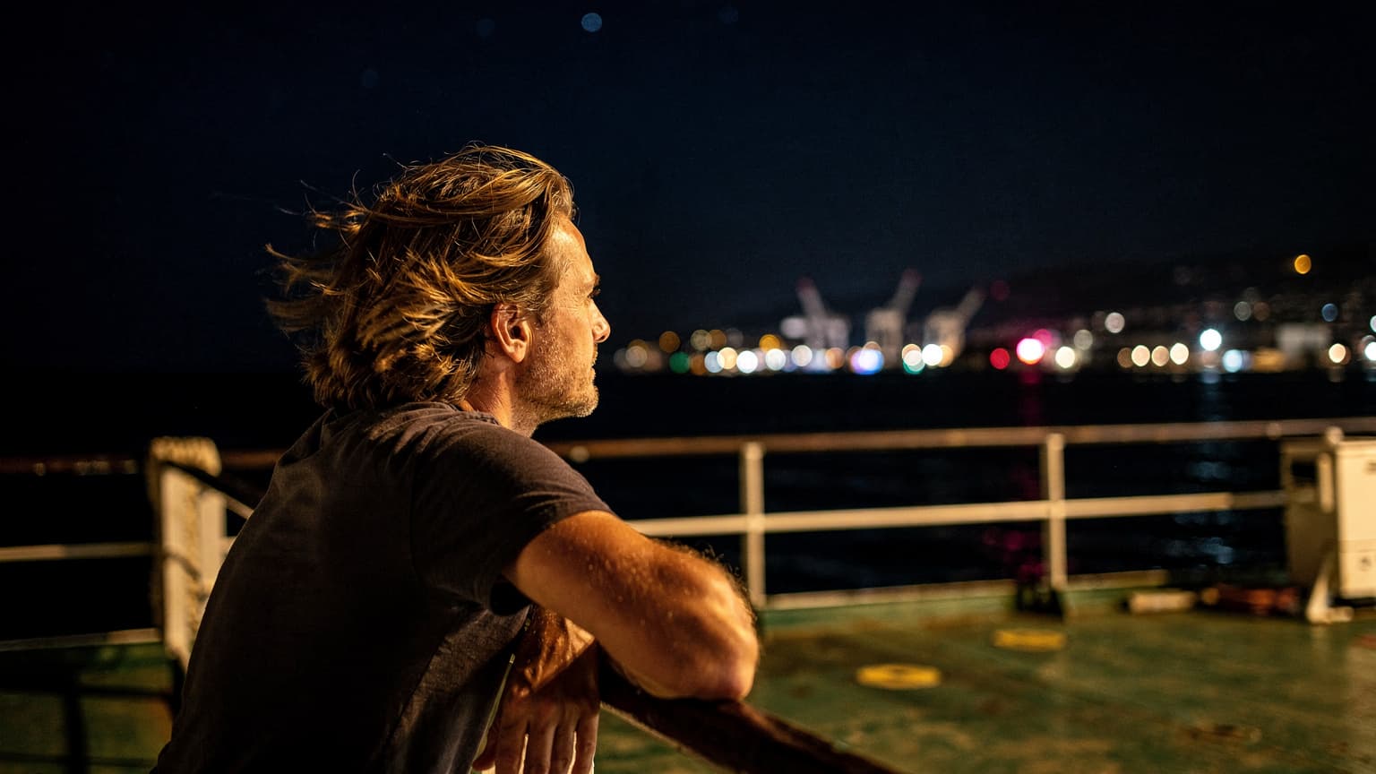 Eine Person lehnt nachts auf dem Deck einer Fähre im Wind, mit beleuchteter Küstenlinie im Hintergrund und dem Schein von Stadtlichtern am Horizont.