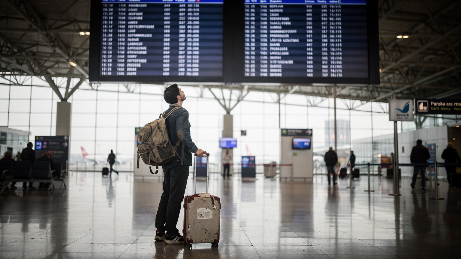 A lone traveler stands at a bustling Charles de Gaulle Airport arrivals hall, gazing at an overhead departures board, luggage neatly at their feet, with the subtle gleam of wet surfaces from a rainy Parisian day.