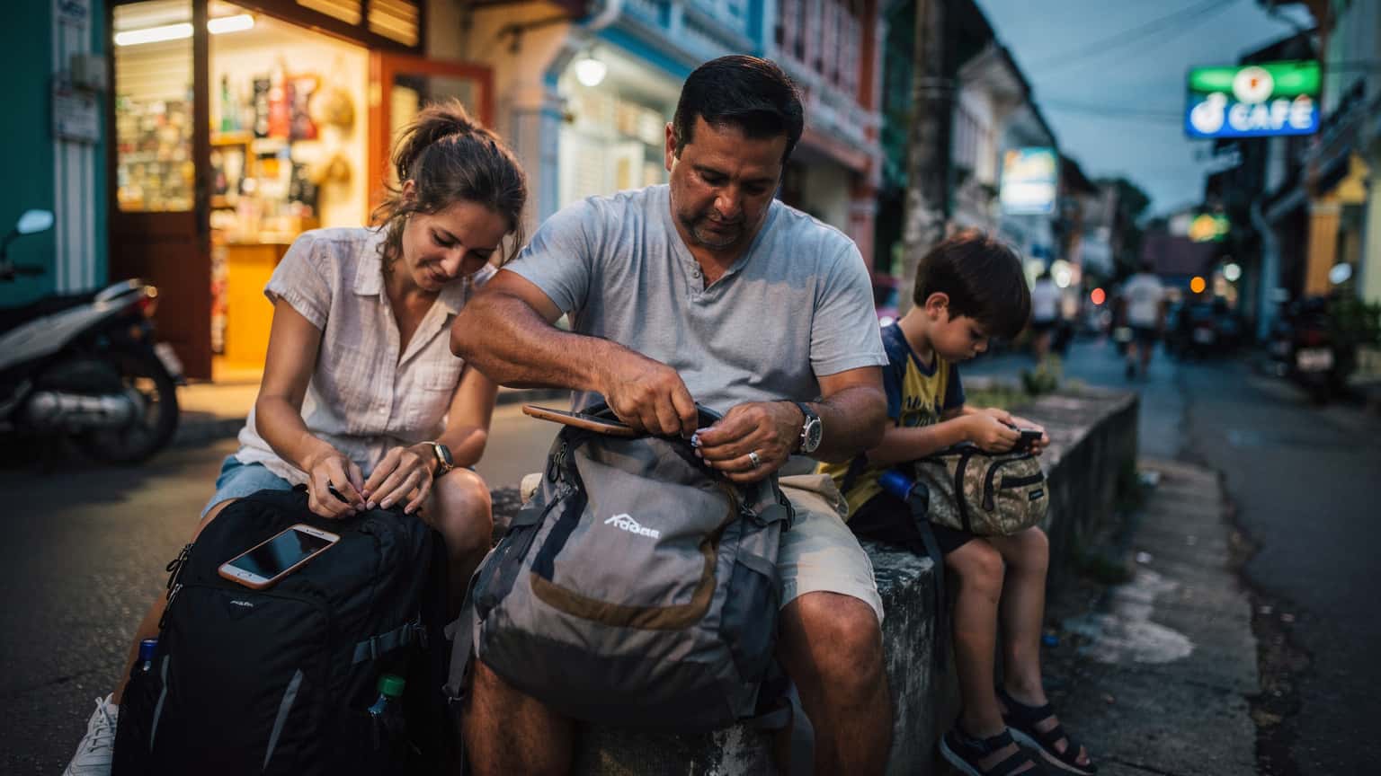 Gia đình ngồi sắp xếp hành lý trên con phố nhỏ ở Phuket lúc hoàng hôn, ánh đèn vàng từ các cửa hàng soi rọi, không ai nhìn vào điện thoại