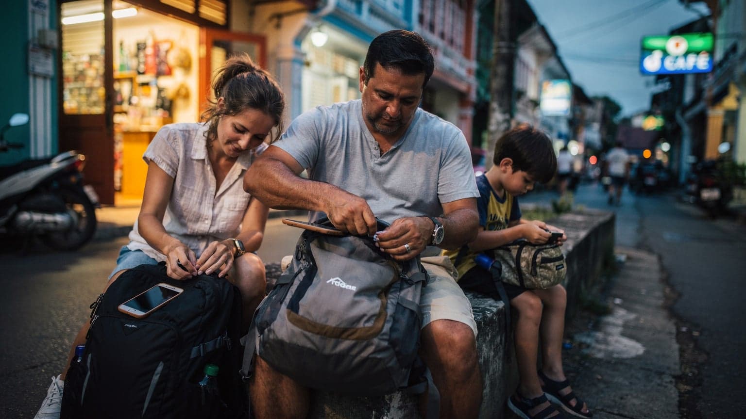 Gia đình ngồi sắp xếp hành lý trên con phố nhỏ ở Phuket lúc hoàng hôn, ánh đèn vàng từ các cửa hàng soi rọi, không ai nhìn vào điện thoại