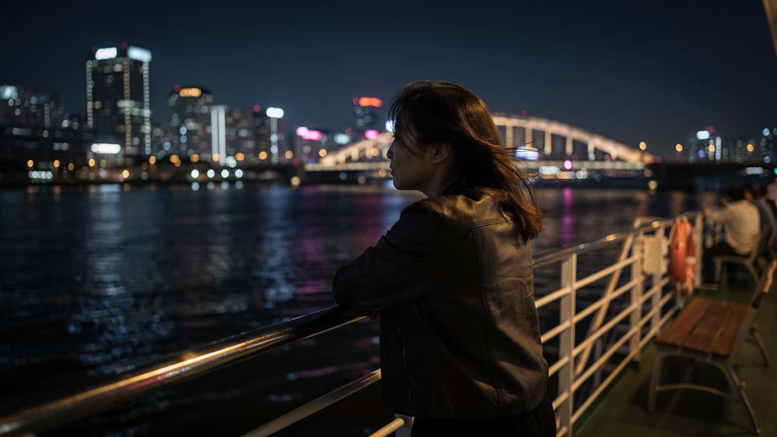 Một người phụ nữ tựa vào lan can phà trên sông Hàn vào buổi tối, gió thổi nhẹ mái tóc, phía sau là những tòa nhà cao tầng với ánh đèn neon rực rỡ phản chiếu trên mặt nước tĩnh lặng.
