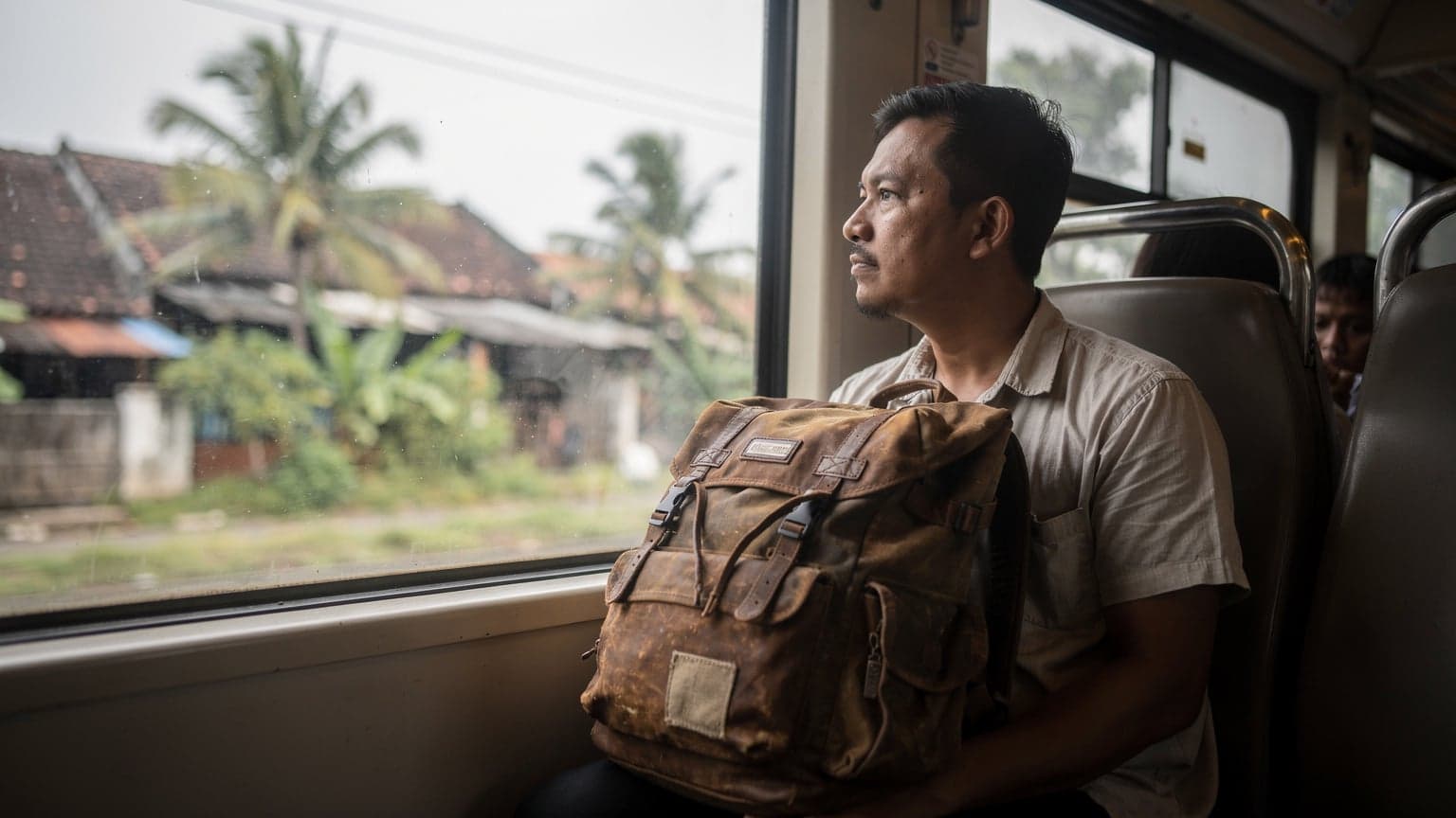 A traveler gazes out of a train window, watching the lush green landscape of suburban Jakarta, Indonesia, pass by in the soft morning light, with their backpack visible on the seat beside them.