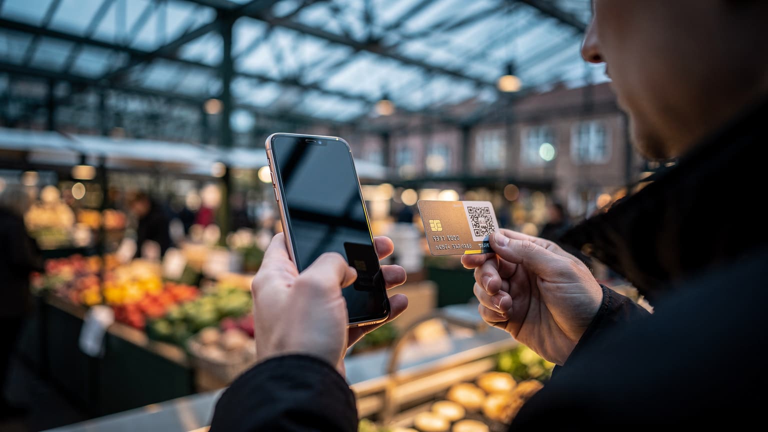 Hænder med en telefon i en overdækket markedsbod i København, skærmen væk fra kameraet, kolde morgentoner