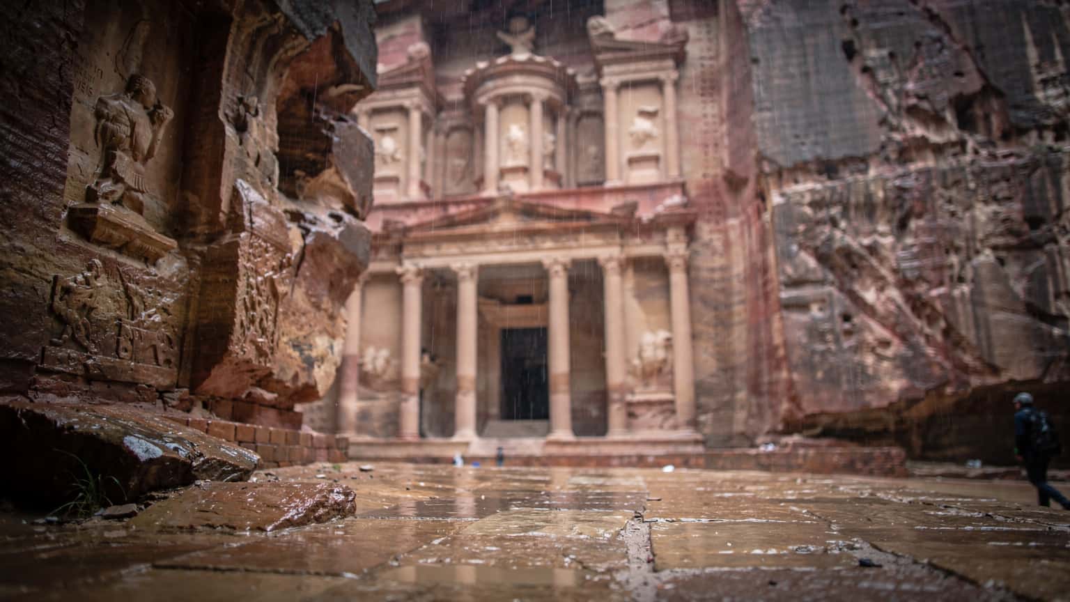 Detail vstupní brány do starověkého města Petra v Jordánsku za deštivého počasí, s mokrými kameny a náznaky architektonických detailů.