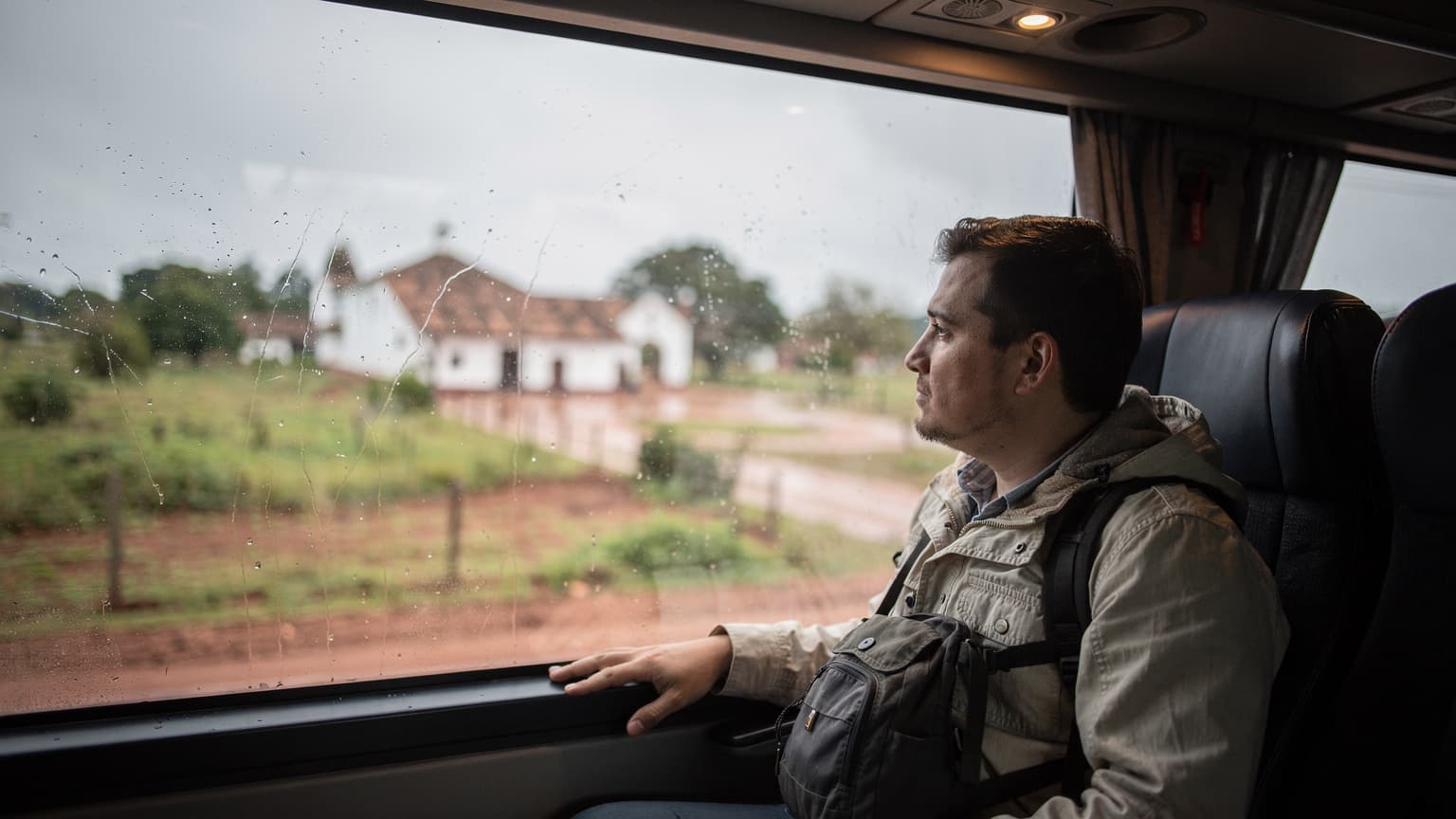 Un viajero mira por la ventana de un autobús en movimiento mientras el paisaje rural de Paraguay, con sus campos verdes y una casa colonial a lo lejos, se difumina por la lluvia.