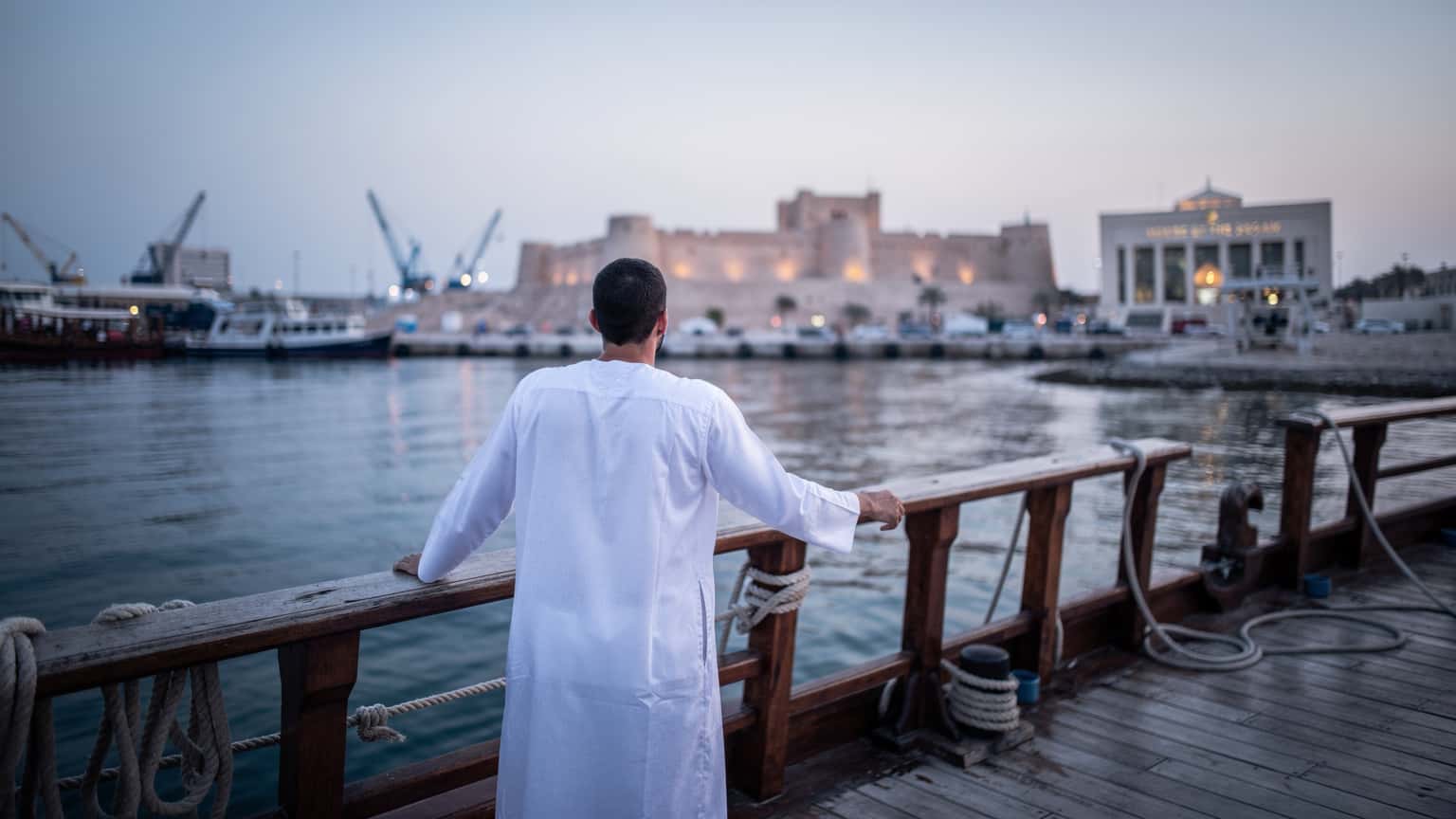 شخص يرتدي ثوبًا تقليديًا عربيًا يتكئ على حاجز مركب في ميناء المنامة، مع مشهد ساحلي لقلعة البحرين وبيت القرآن في خلفية زرقاء باردة لشروق الشمس.