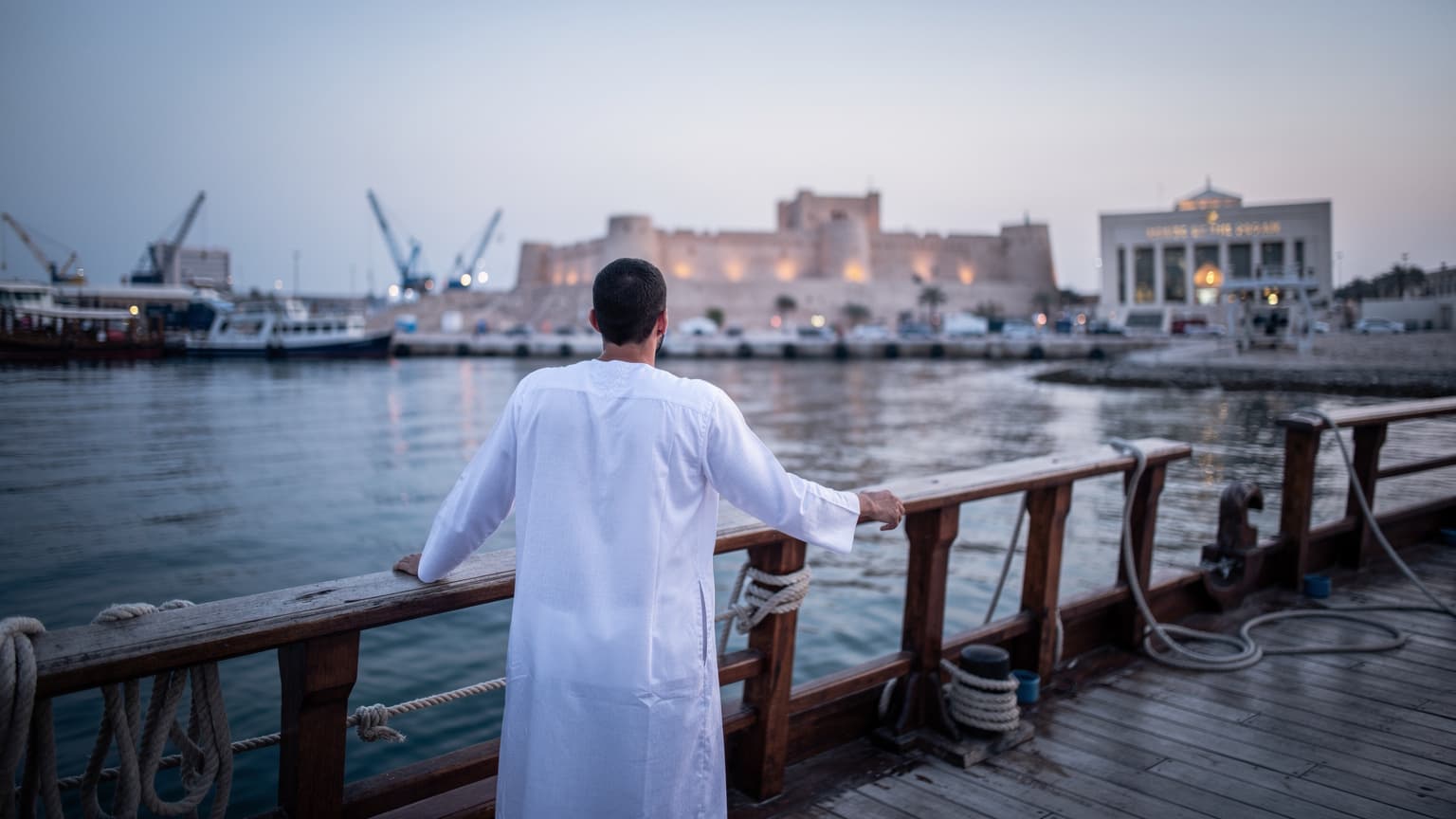 شخص يرتدي ثوبًا تقليديًا عربيًا يتكئ على حاجز مركب في ميناء المنامة، مع مشهد ساحلي لقلعة البحرين وبيت القرآن في خلفية زرقاء باردة لشروق الشمس.