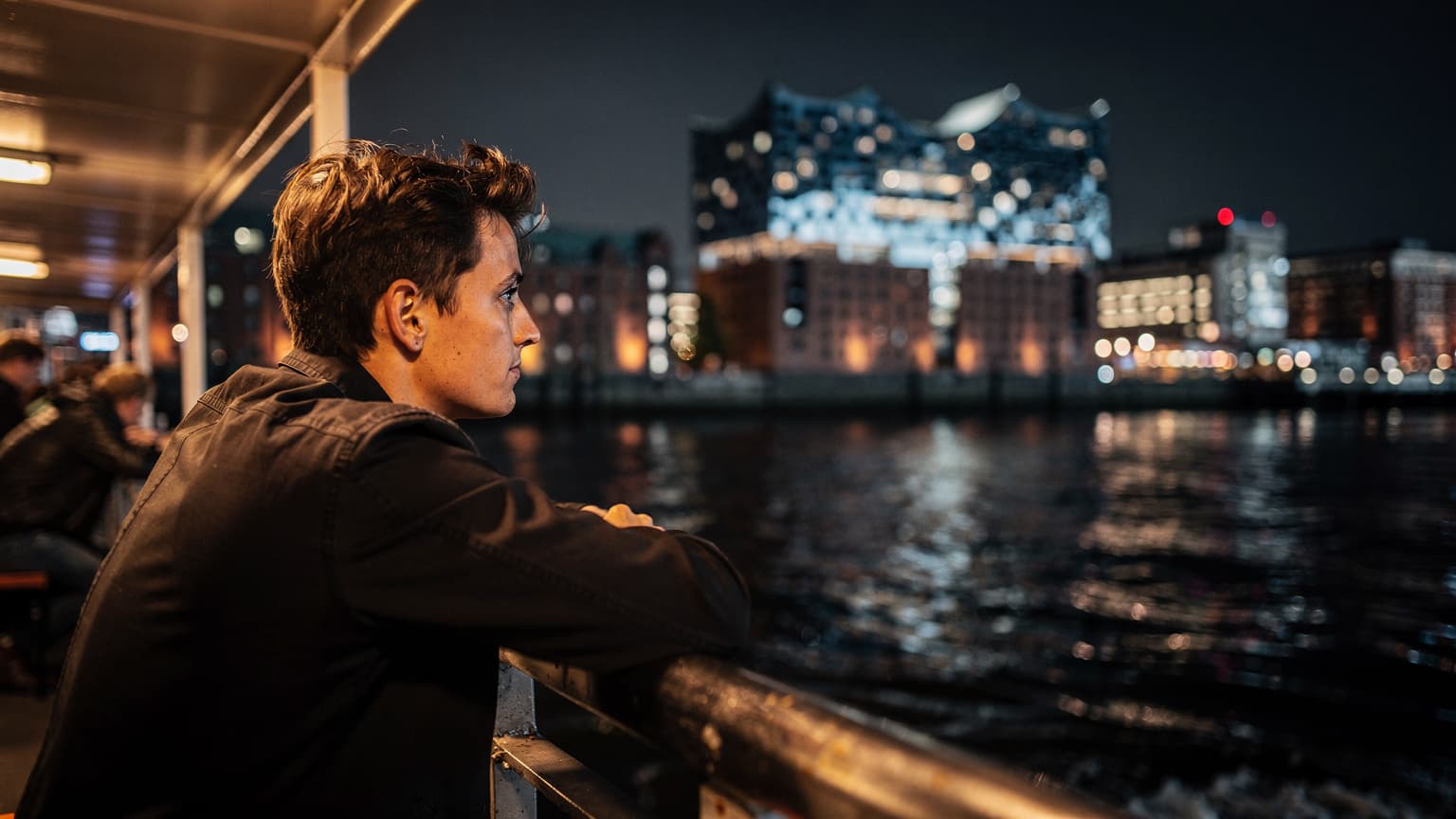 Eine Person lehnt nachts auf dem Deck einer Fähre im Hamburger Hafen, der Wind weht durch ihr Haar, im Hintergrund sind die beleuchtete Elbphilharmonie und die Docks zu sehen.