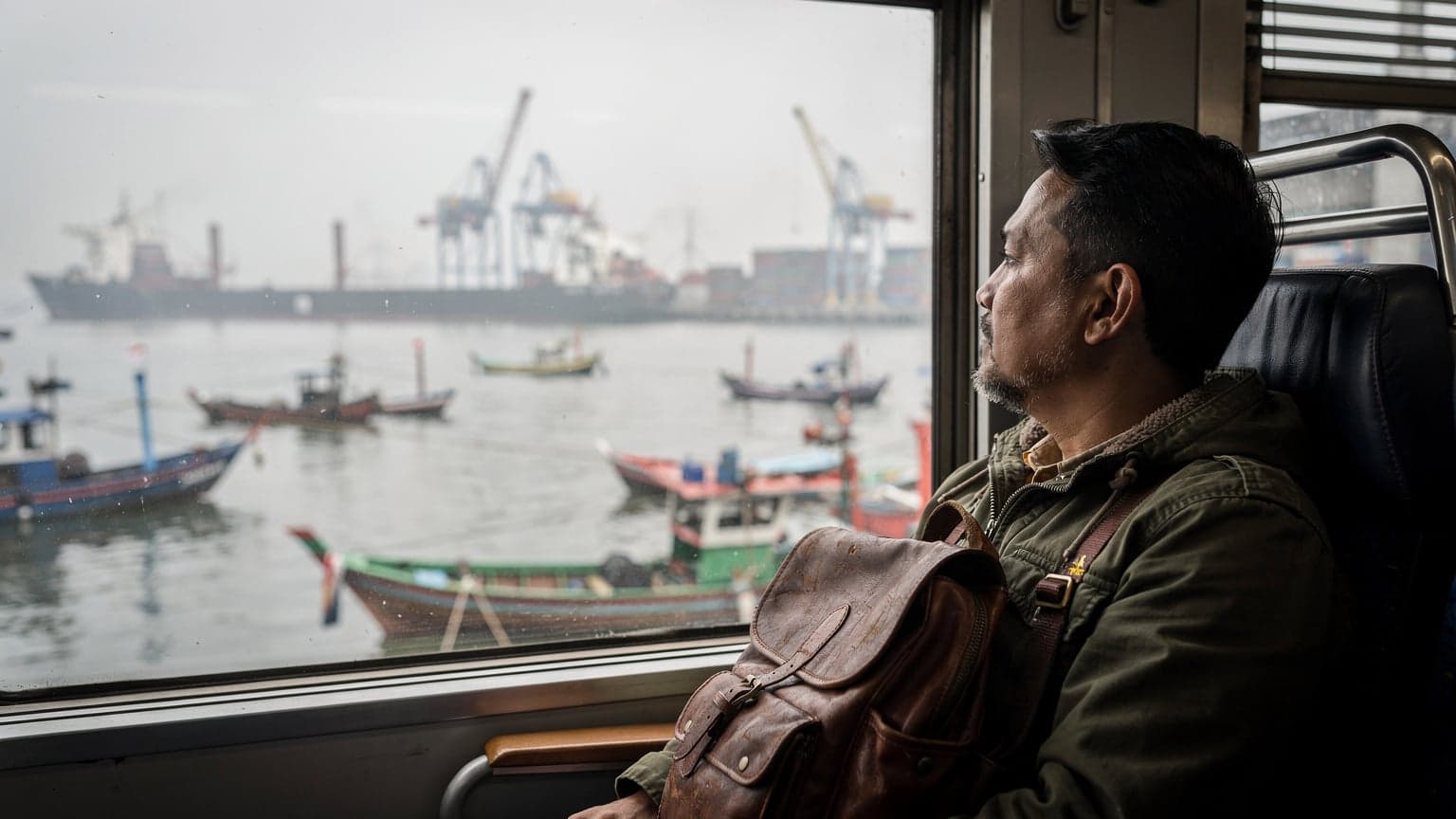 En reisende ser ut av vinduet på et tog som kjører langs havnen i Jakarta, med en ryggsekk på setet ved siden av seg.