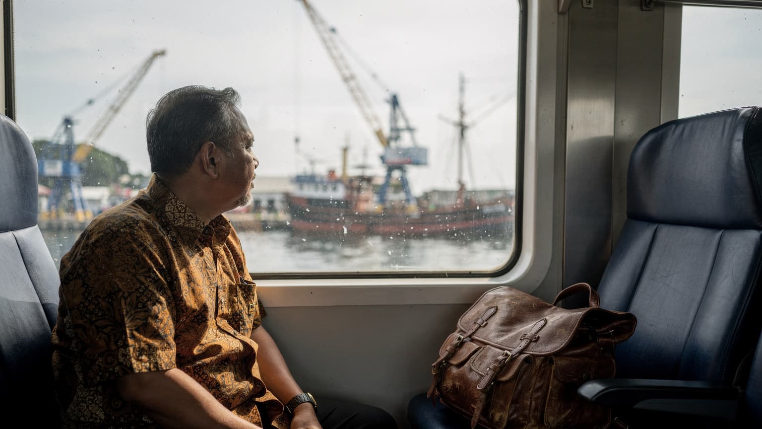 Penumpang KA Commuter Line melihat pelabuhan Sunda Kelapa dari jendela kereta, tas di kursi sebelahnya, cahaya pagi mendung