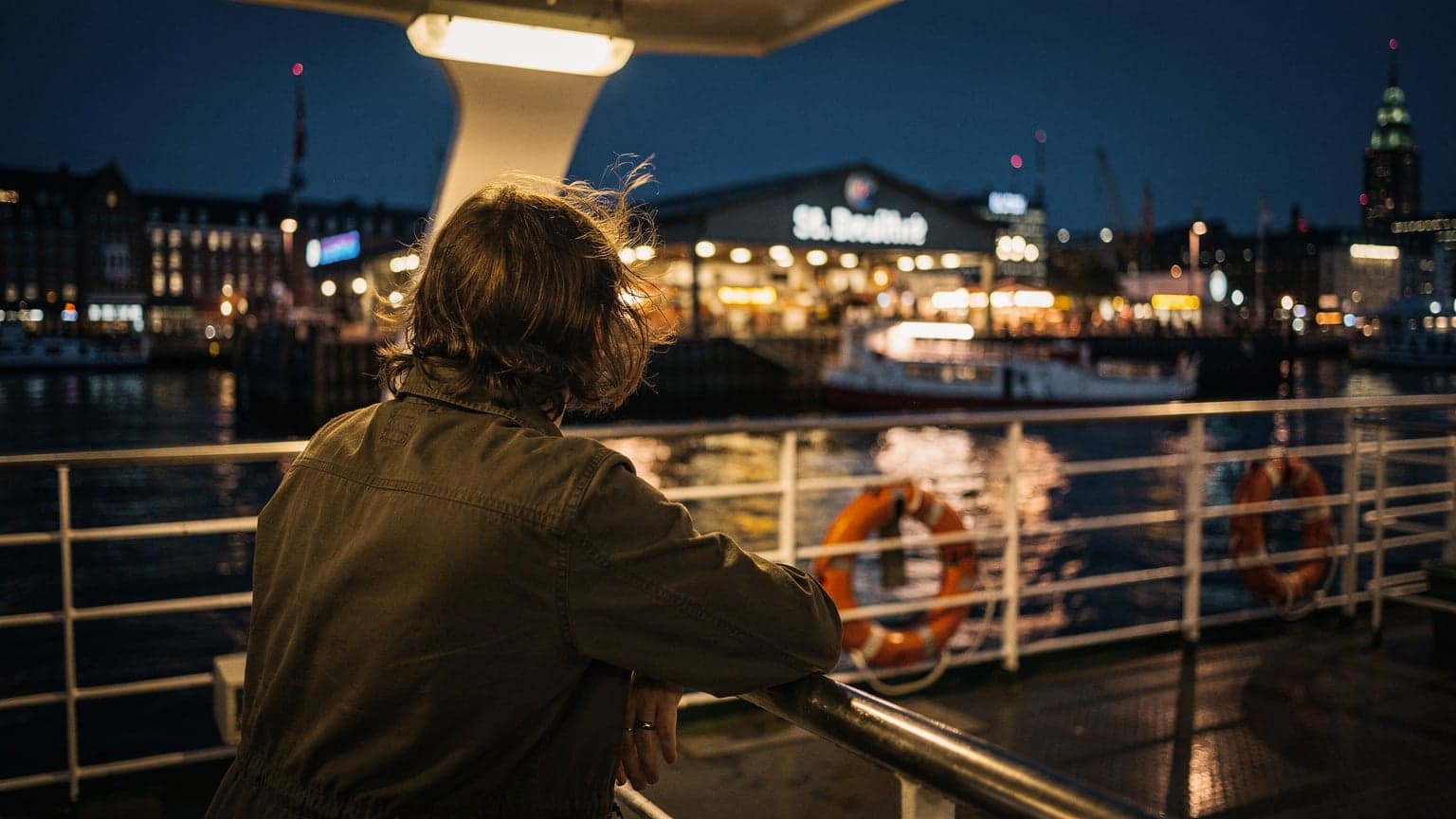 Eine Person lehnt sich an das Geländer einer Fähre in Hamburg bei Nacht, mit dem beleuchteten Fischmarkt im Hintergrund und dem Wind in den Haaren.