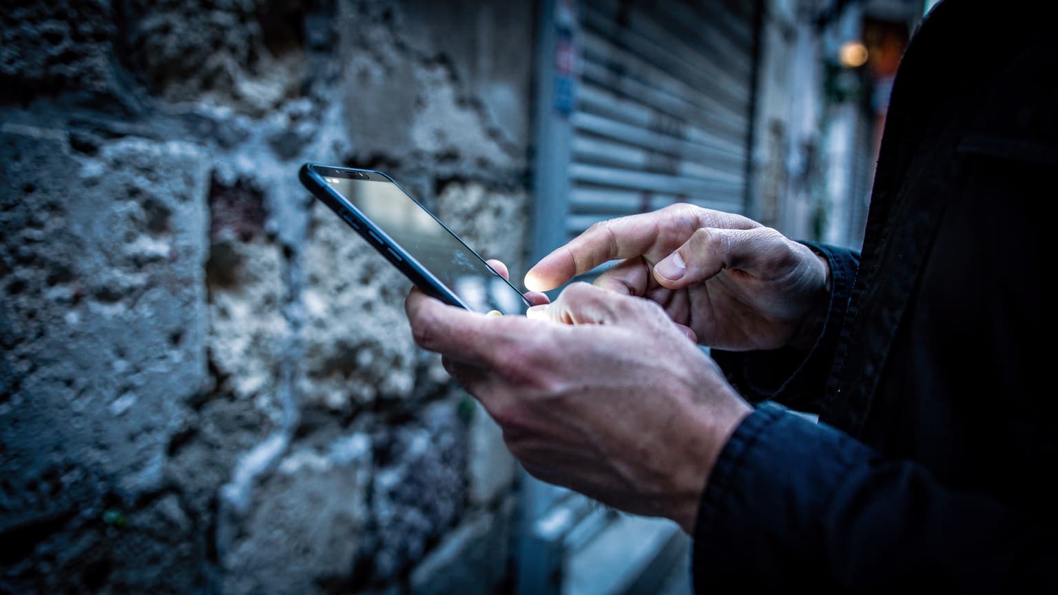 Karaköy'de, arka sokaklardan birinde, soğuk mavi tonlarda sabah ışığında bir kişinin elleri akıllı telefon ekranına odaklanmış, cihazın arka yüzü kameraya dönük.