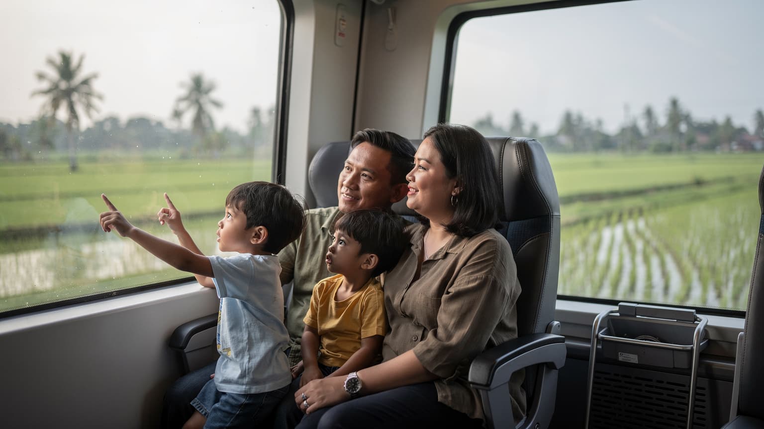 A family on a train in Indonesia, a child looking out the window at lush green rice paddies, their parents relaxed in the seats beside them.