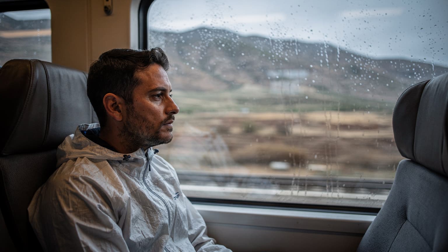 Viajante a observar a paisagem marroquina através da janela de um comboio, saco ao lado, sob luz cinzenta de chuva, na estação de Tanger.
