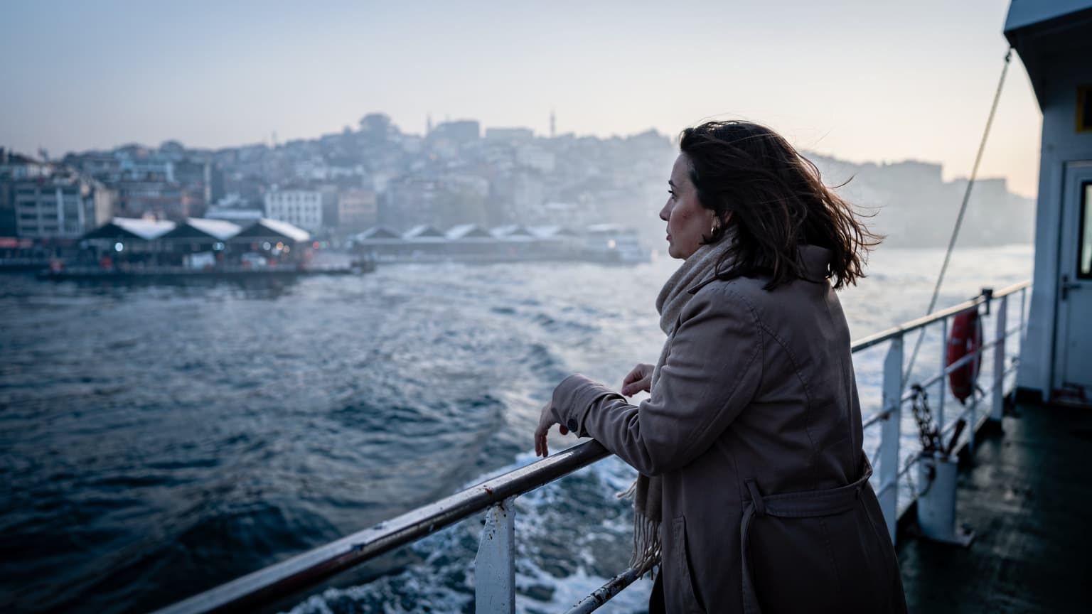 Bir kadın, İstanbul'daki Sabiha Gökçen Havalimanı'ndan kalkan bir feribotun güvertesinde, rüzgarda saçları uçuşurken, arkasında Anadolu yakasının sahil şeridi uzanıyor.