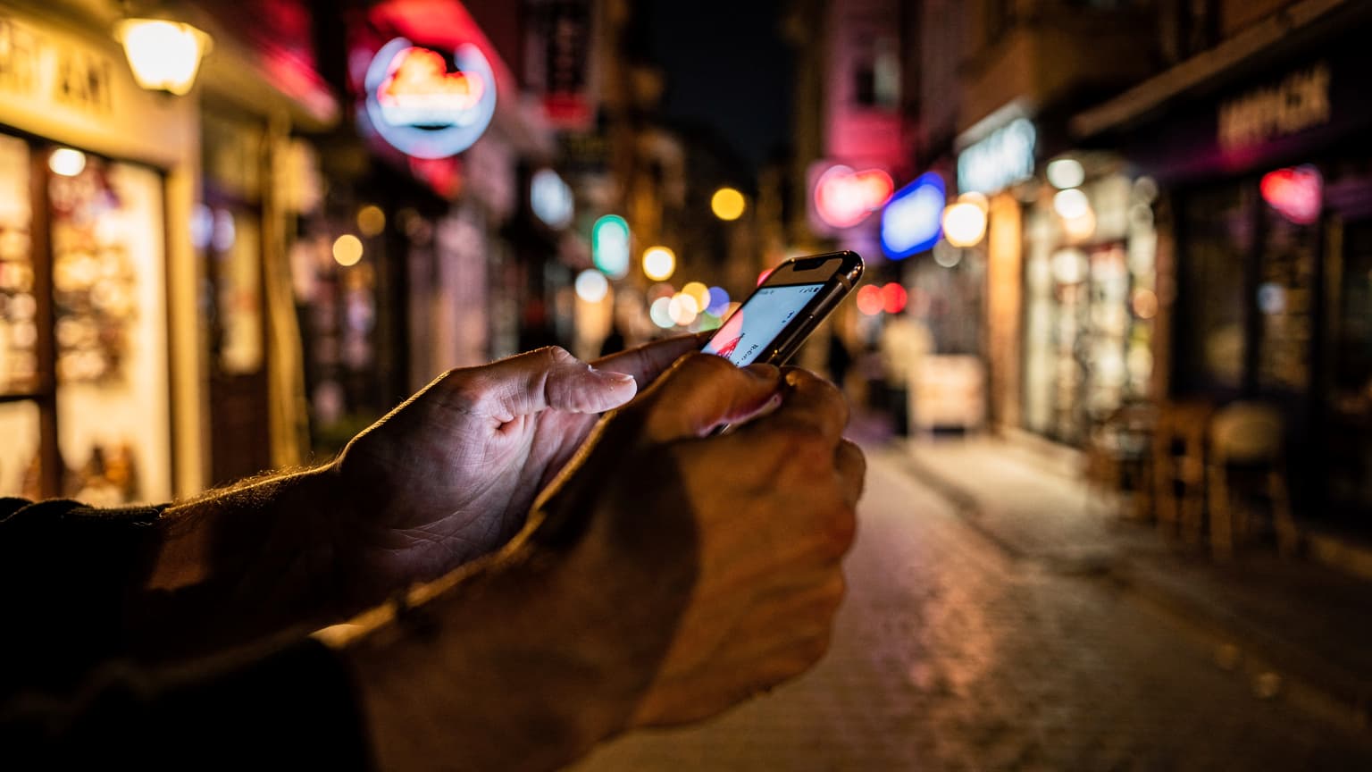 Close-up of a traveler's hands holding a smartphone, with the screen turned away, silhouetted against the warm glow of neon signs and tungsten streetlights on a bustling back street in Istanbul's Beyoğlu district at night.