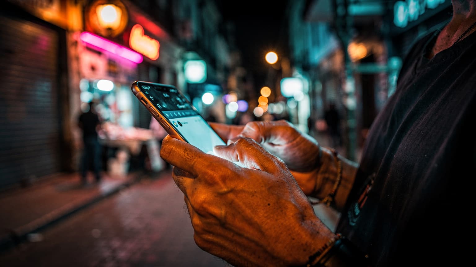 Close-up of hands typing on a smartphone keyboard in a dimly lit, vibrant back street market in Buenos Aires, Argentina, at night.