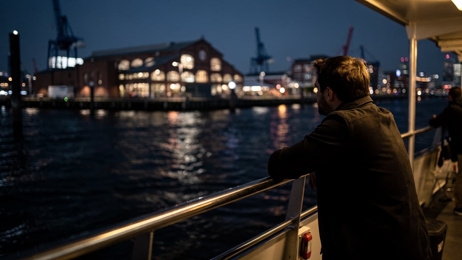 Eine Person lehnt sich an das Geländer einer Fähre im Hamburger Hafen bei Nacht, im Hintergrund schimmern die Lichter des Fischmarktes und der Elbe.