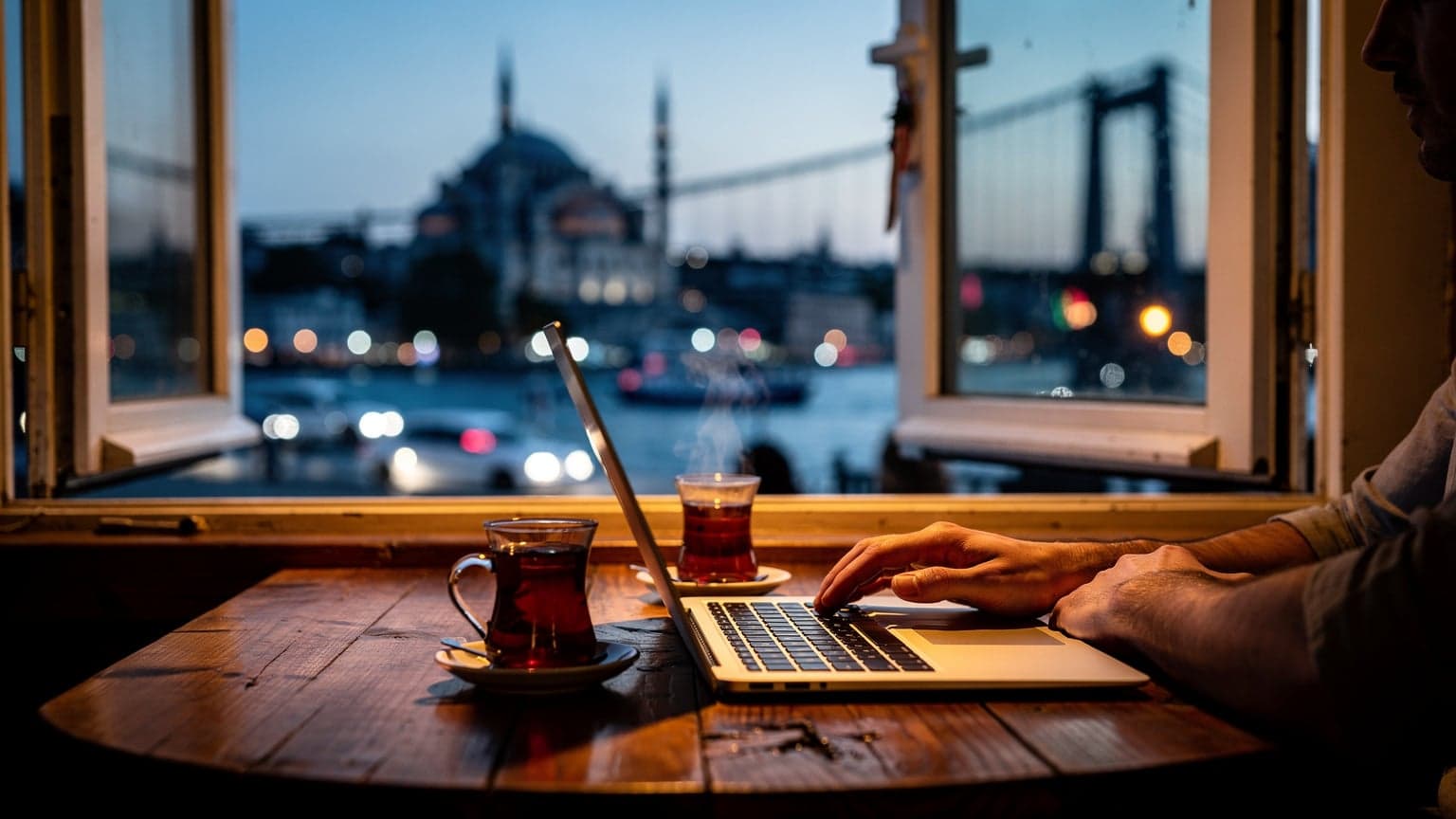 Pohľad z okna kaviarne v istanbulskej štvrti Karaköy počas modrej hodiny, na stole laptop a čaj, za oknom mešita a Bospor