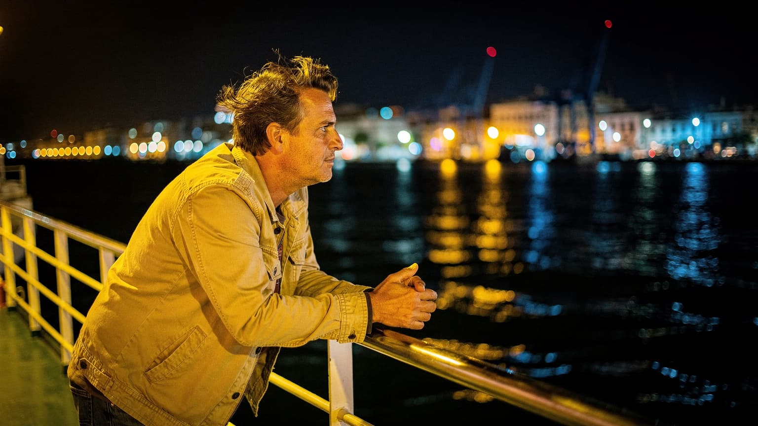 Un voyageur penché sur le bastingage d'un ferry dans le port de La Goulette, la nuit, avec les lumières de Tunis en arrière-plan et le vent dans les cheveux
