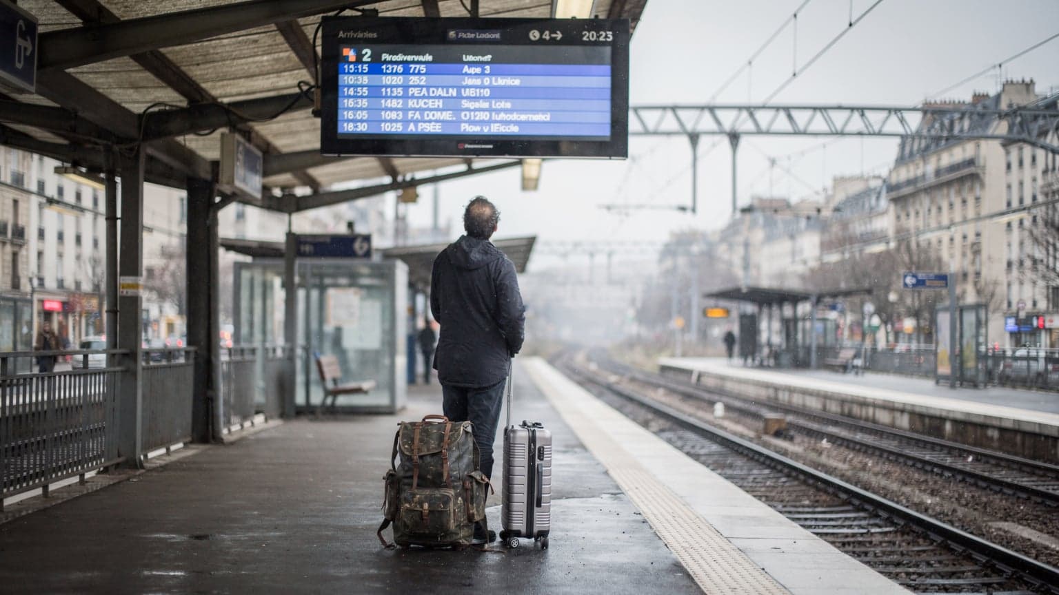 A lone traveler stands on a damp Parisian suburban rail platform, gazing up at a large, illuminated arrivals and departures board in the distance, luggage at their feet, as rain streaks down the station's glass roof.