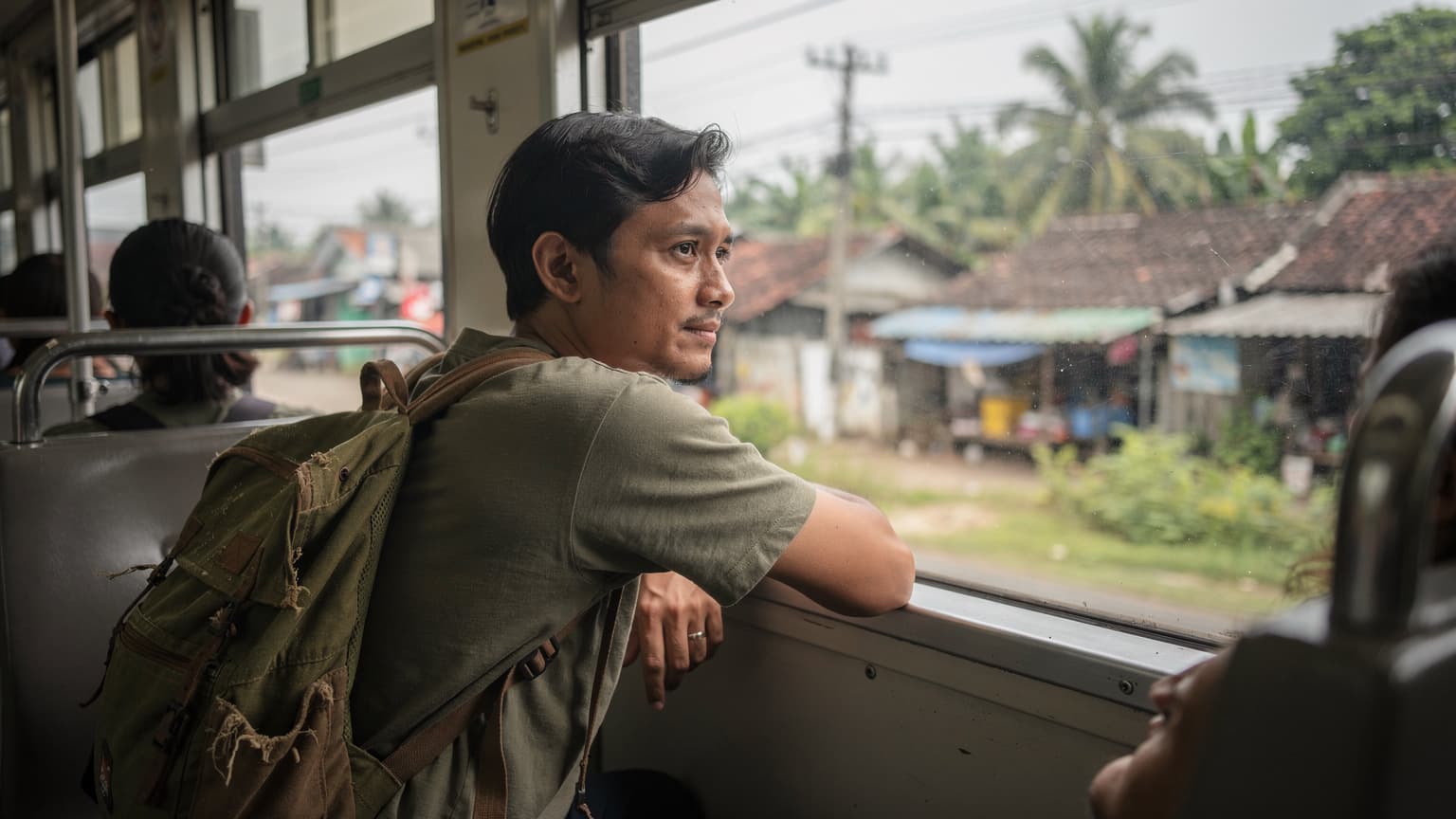 A traveler gazes out a train window in Jakarta's suburbs during a soft, overcast morning, with their backpack resting on the seat beside them.