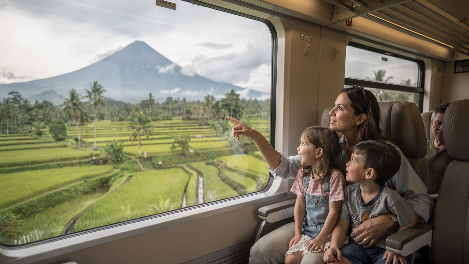 A family watching the Balinese landscape, rich with rice paddies and volcanic peaks, slide past the window of a comfortable, air-conditioned train carriage during a mid-morning journey.