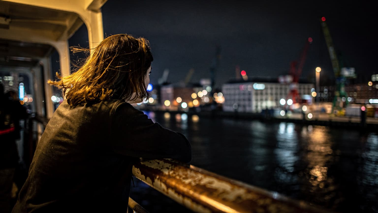 Eine Person lehnt nachts auf der Reling einer Fähre im Hamburger Hafen, der Wind zerzaust ihr Haar, im Hintergrund sind die beleuchteten Docks und Gebäude des Fischmarktes zu sehen.