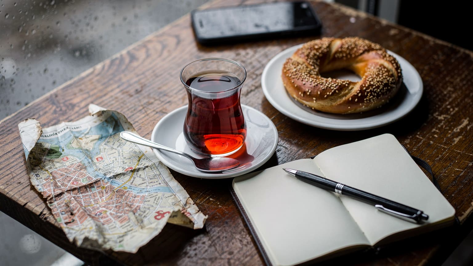 Vista dall'alto di un tavolino di un caffè a Kadıköy, Istanbul, con un tè turco in un bicchiere a tulipano, un piatto di simit e una mappa cartacea spiegazzata, luce piovosa grigia dalla finestra