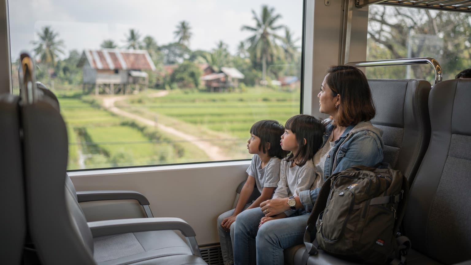 A family with two children, aged around 8 and 10, looking out a train window at a lush green Indonesian landscape, with traditional houses visible in the distance, during a soft overcast mid-morning.