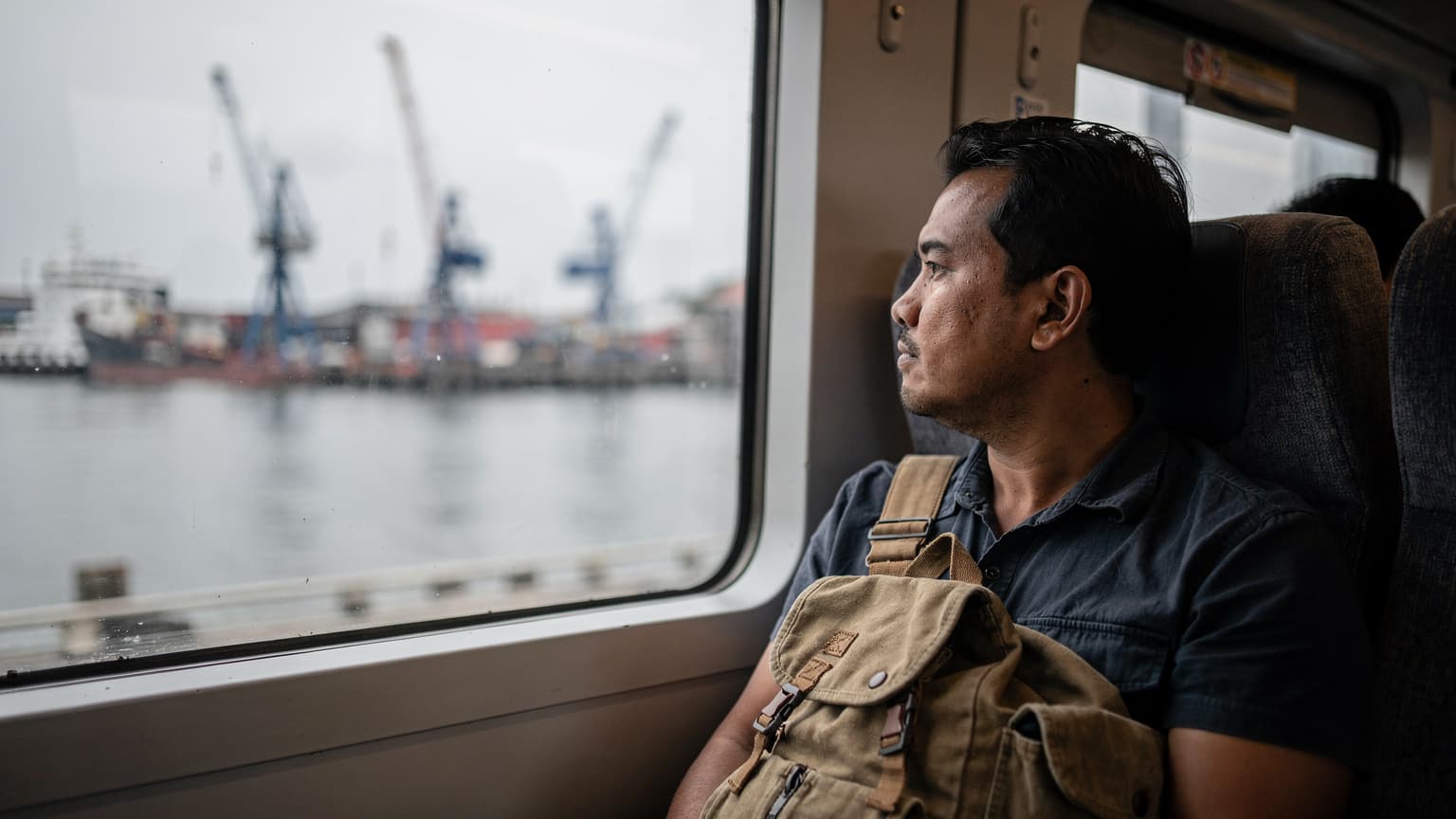 Pria dengan tas ransel melihat keluar jendela kereta commuter di pagi hari, melintasi pelabuhan Sunda Kelapa, Jakarta