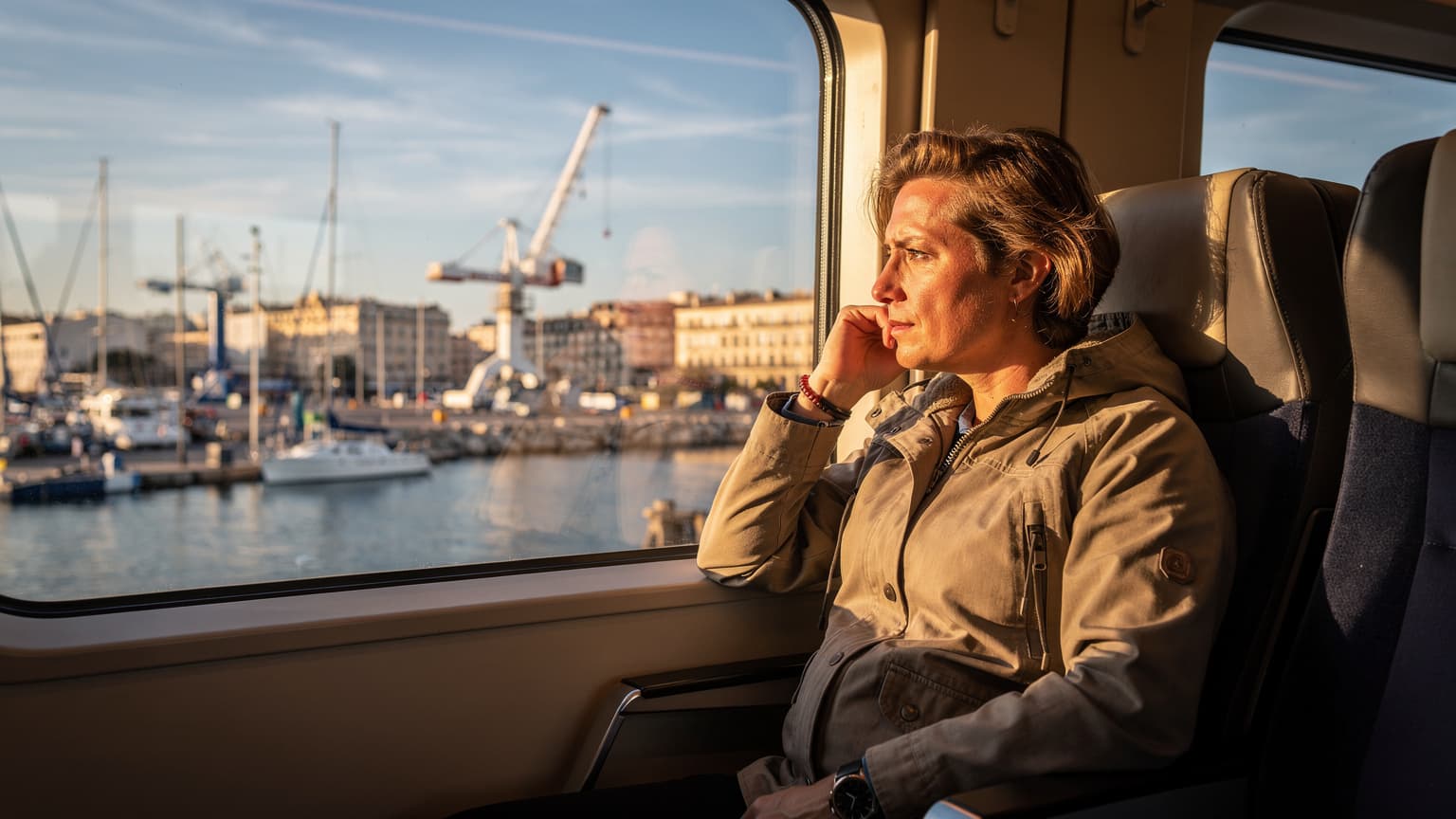 A traveler gazes out a train window at a bustling harbor in Marseille during late afternoon, a backpack resting on the seat beside them.