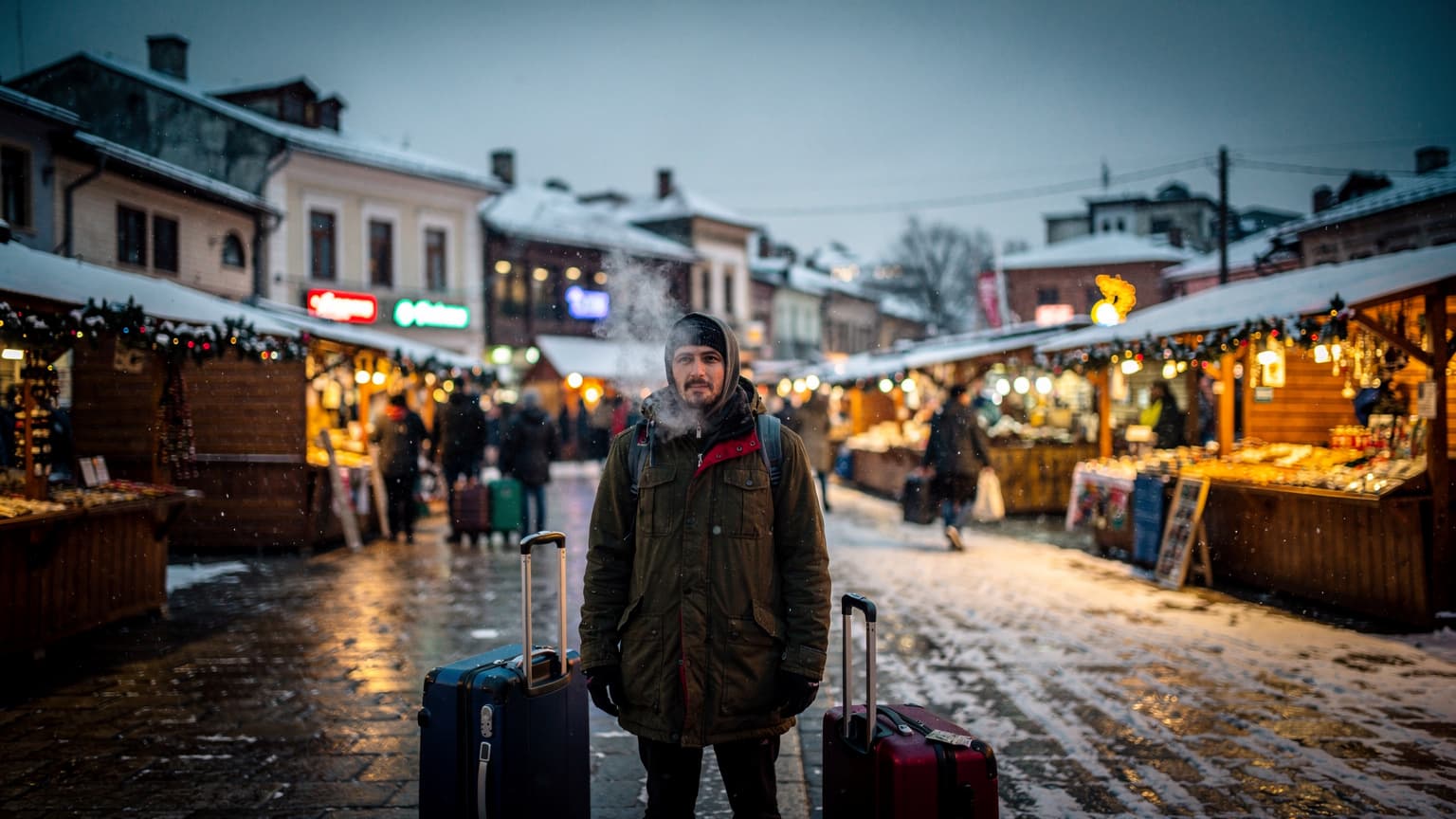 Eine Person wartet auf einem verschneiten Weihnachtsmarkt in Sarajevo, umgeben von gedämpftem Licht von Ständen und Laternen. Sie steht mit einem aufrechten Koffer da, der Blick in die Ferne gerichtet, während Neon- und Wolframlicht die Szene beleuchten.