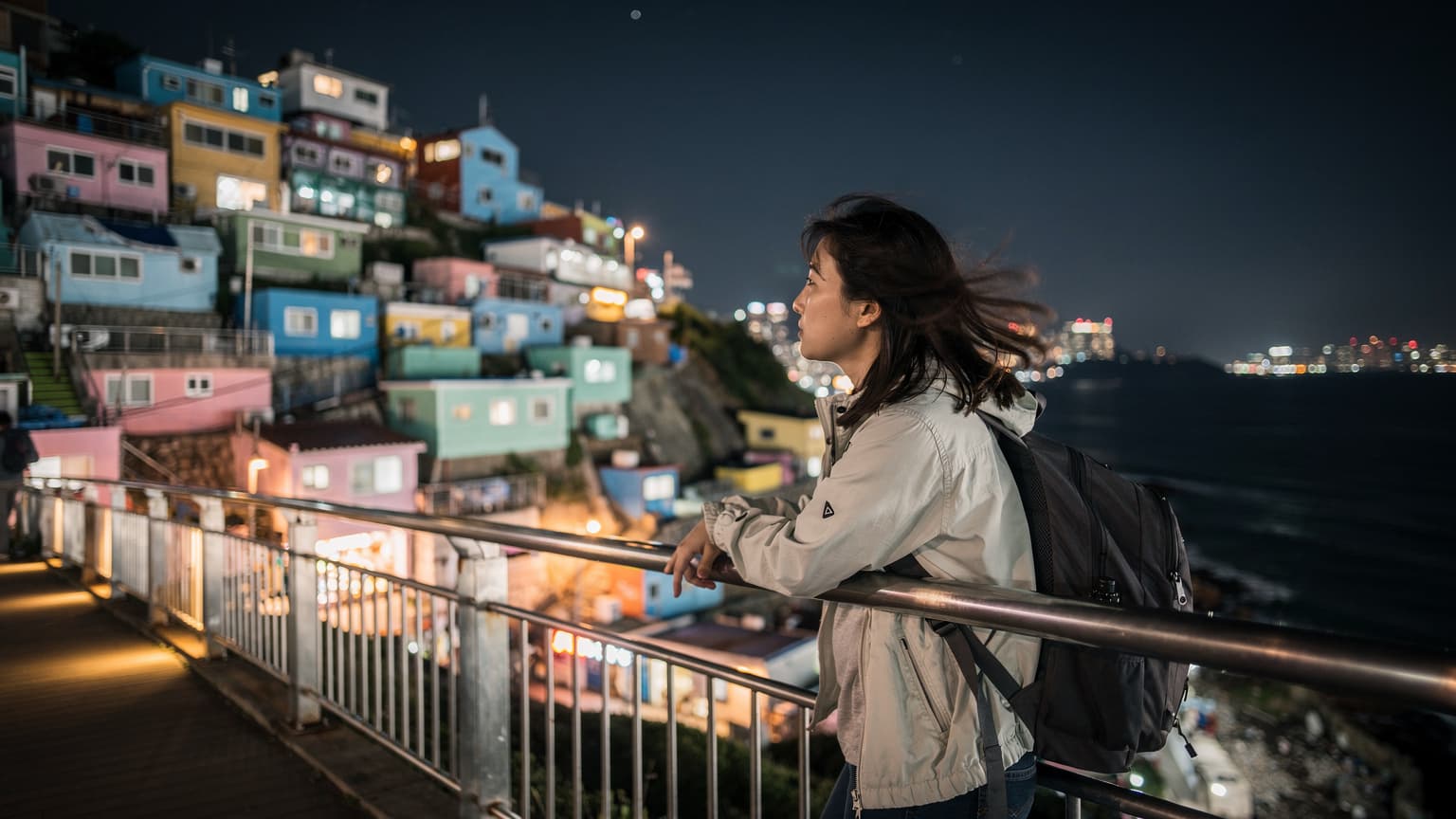 夜幕下的釜山甘川洞文化村,一位游客扶着海边步道的栏杆,回望色彩斑斓的村落灯火,海风吹拂。