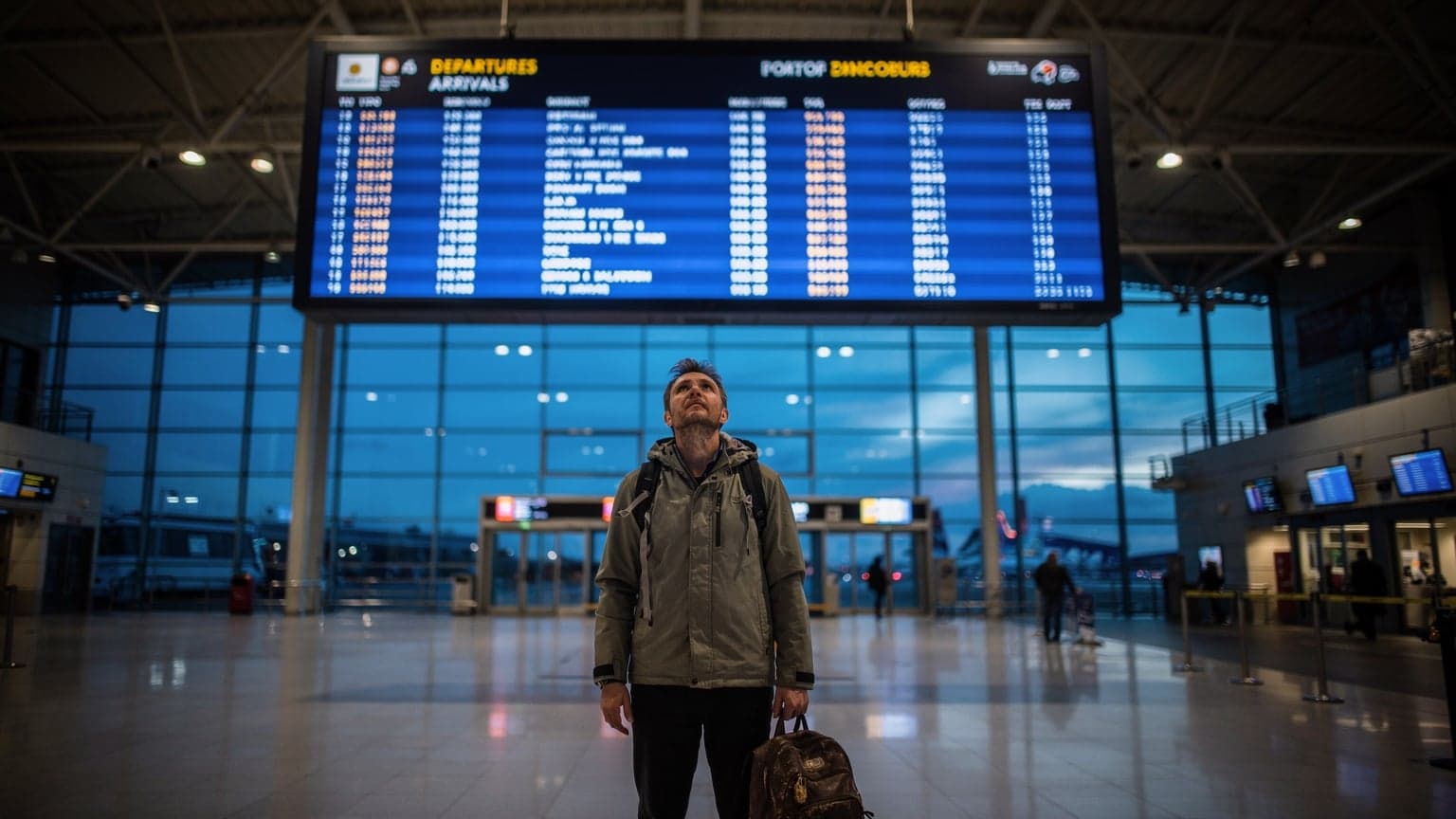 A traveler, with luggage at their feet, looks up at a large, illuminated arrivals and departures board in the blue hour just after sunset at the Port of Dover's ferry terminal.