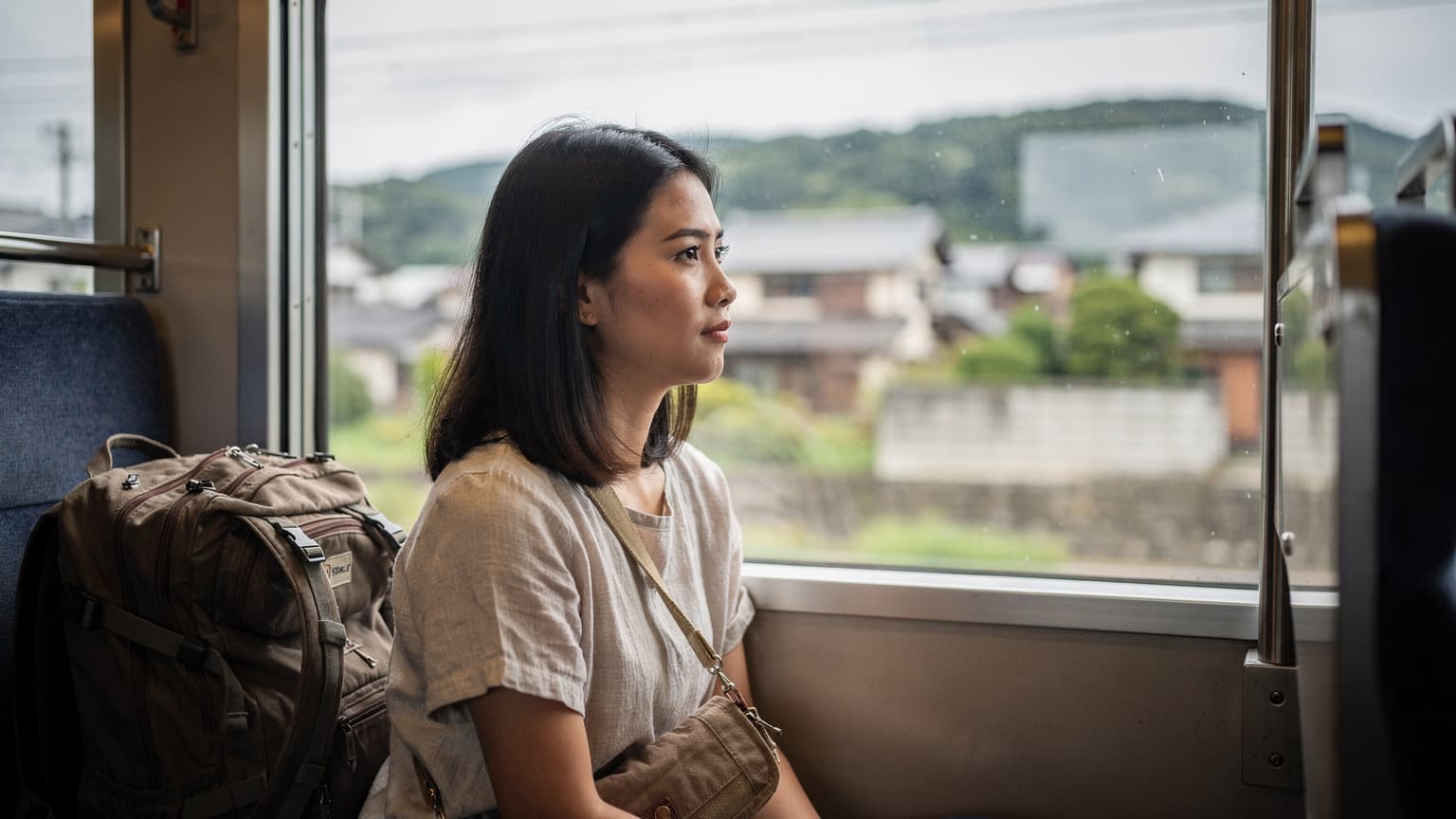 นักเดินทางหญิงนั่งรถไฟชานเมืองมองทิวทัศน์ผ่านหน้าต่าง กระเป๋าเป้ใบเล็กวางบนที่นั่งข้างๆ แสงกลางวันสลัวยามเช้า บรรยากาศเรียลเหมือนภาพสารคดี