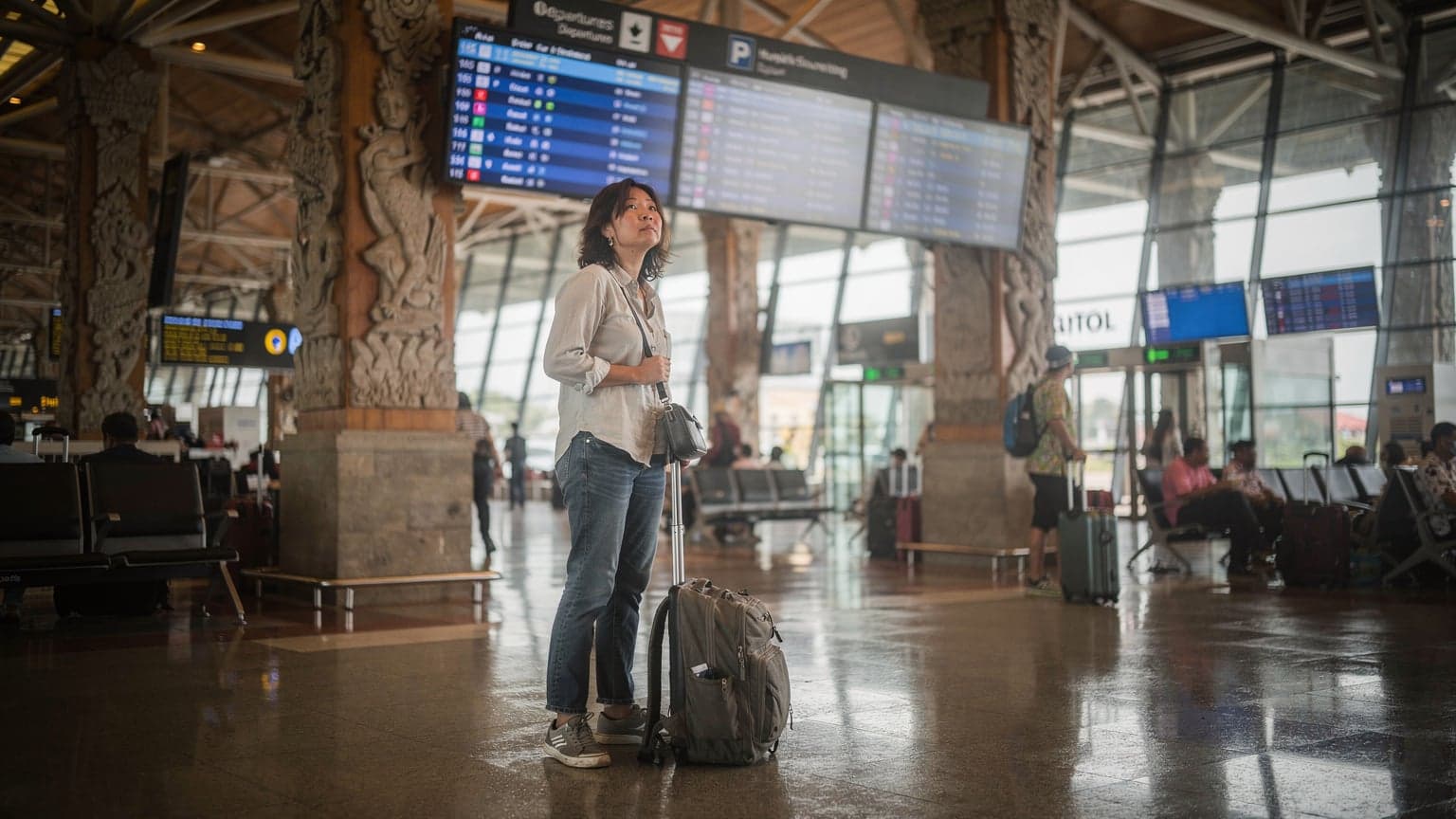 A traveler stands in the arrivals hall of Ngurah Rai International Airport in Denpasar, Bali, looking at an overhead departures board with their luggage at their feet, under grey rainy light.