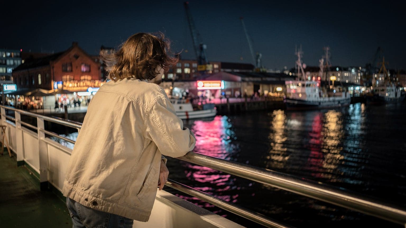 Eine Person lehnt auf der Reling einer Fähre im Hamburger Hafen bei Nacht, im Hintergrund die erleuchteten Docks des Fischmarktes.