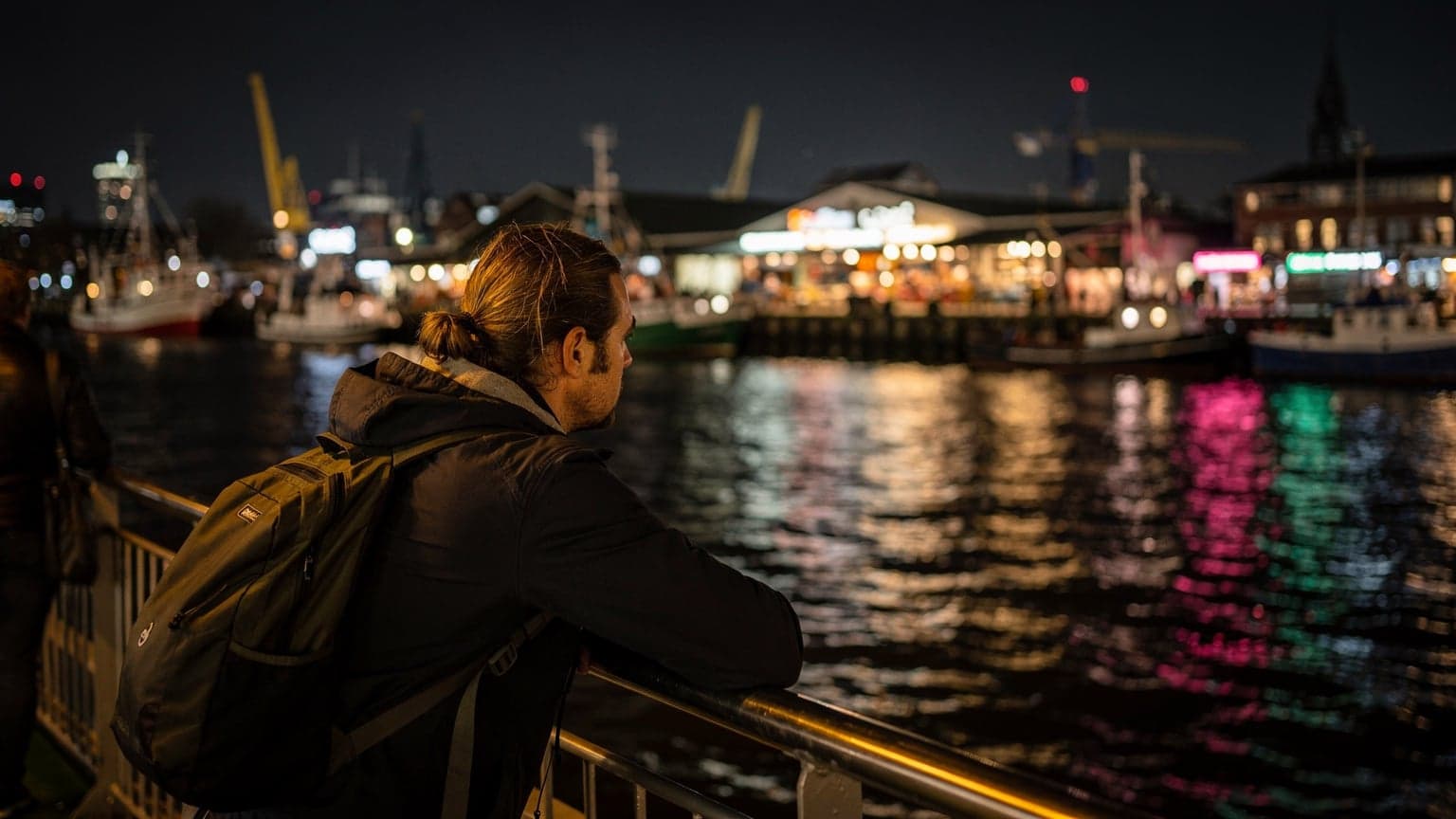 Eine Person lehnt an der Reling einer Fähre bei Nacht, der Wind fährt durch ihr Haar, im Hintergrund sind die Lichter eines Hamburger Fischmarktes an der Küste zu sehen.