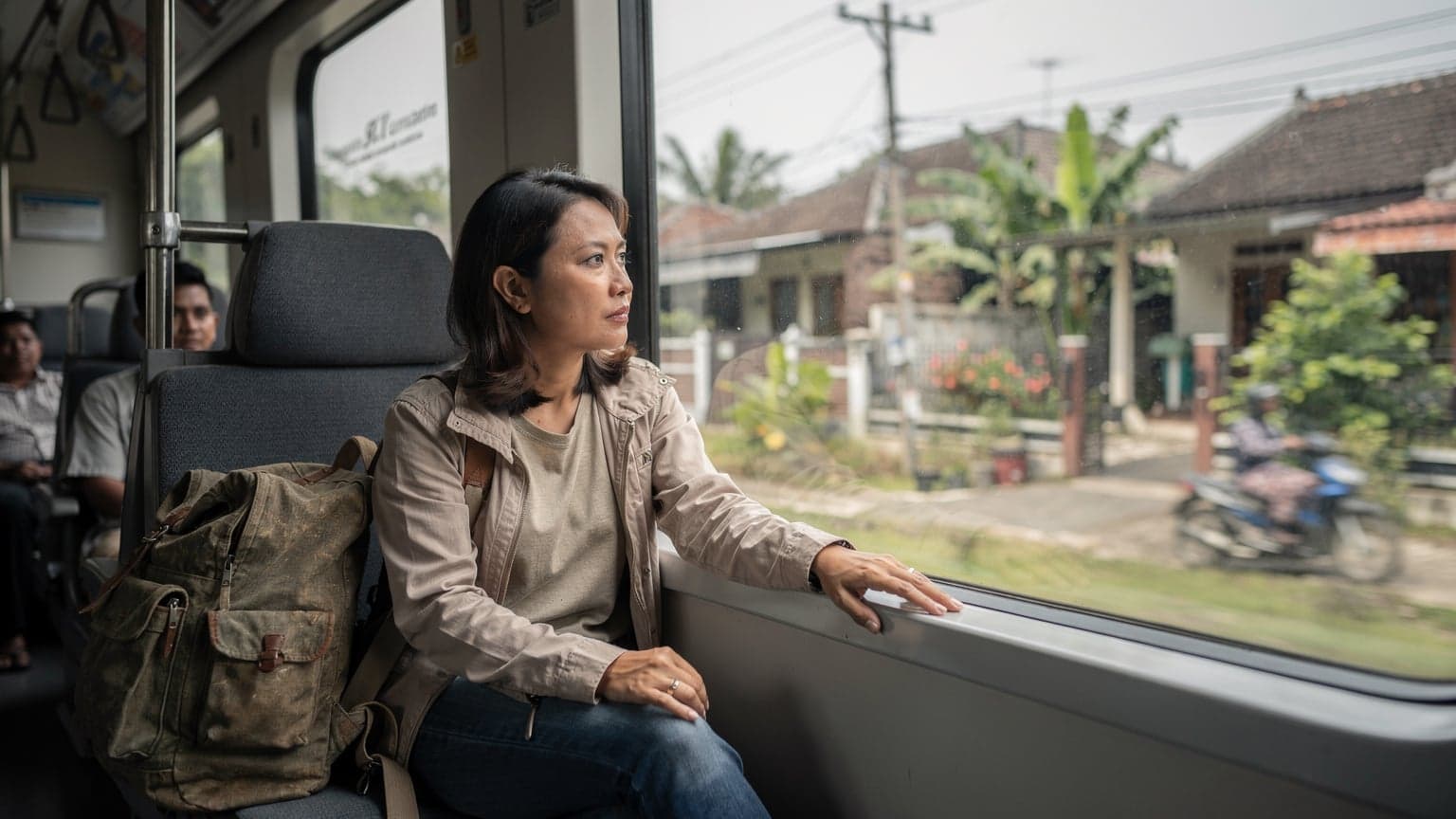 A traveler gazes out a train window at a lush green Indonesian landscape, their backpack resting on the seat beside them, the morning light softly illuminating the carriage.