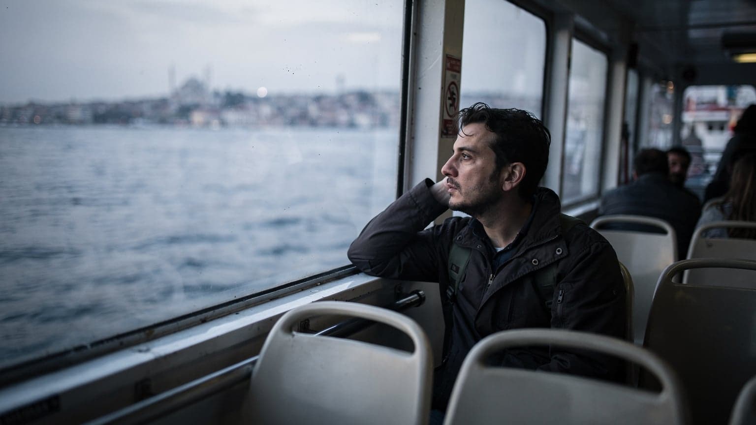 Marmara Denizi'nde seyreden bir vapurda, yorgun argın ama umutlu bir gezginin pencereden dışarı baktığı soğuk mavi tonlarda bir sabah manzarası. Bavulu yanında duruyor, yüzünde hafif bir tebessüm var.