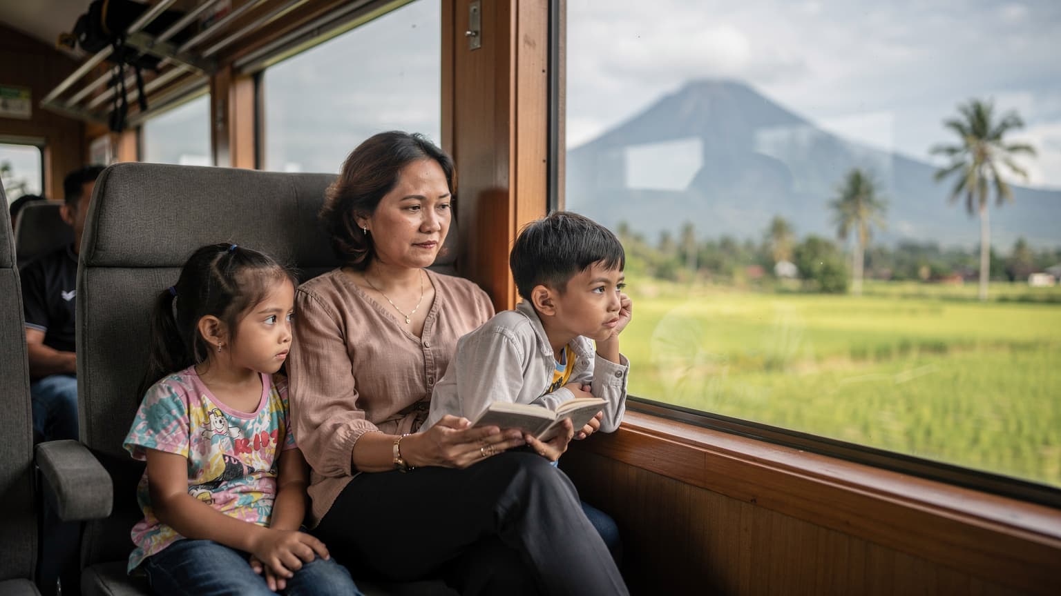 A family traveling on an Indonesian train, a child looking out the window at passing rice paddies and distant volcanic mountains, reflecting the interior of the carriage.