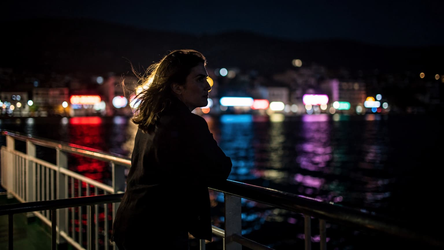 Antalya'da bir sahil kasabasında, gece vakti feribot güvertesinde rüzgarda saçları dalgalanan bir yolcu, arkasında neon ışıklı kıyı şeridi.