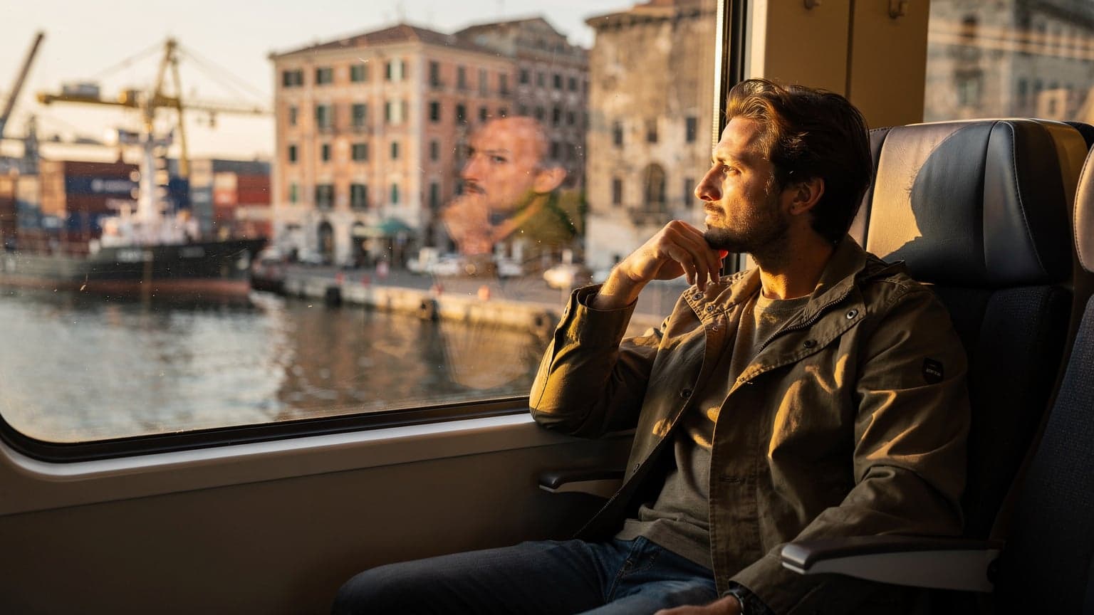 A traveler gazes out of a train window at a bustling harbour front in Genoa, Italy, with their backpack on the seat next to them, late afternoon light casting long shadows.