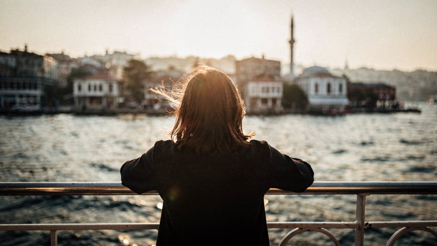 İstanbul'da bir Marmara vapuru güvertesinde, rüzgarda saçları dalgalanan bir yolcu, arkasında tarihi sahil şeridi, altın saat ışığında.