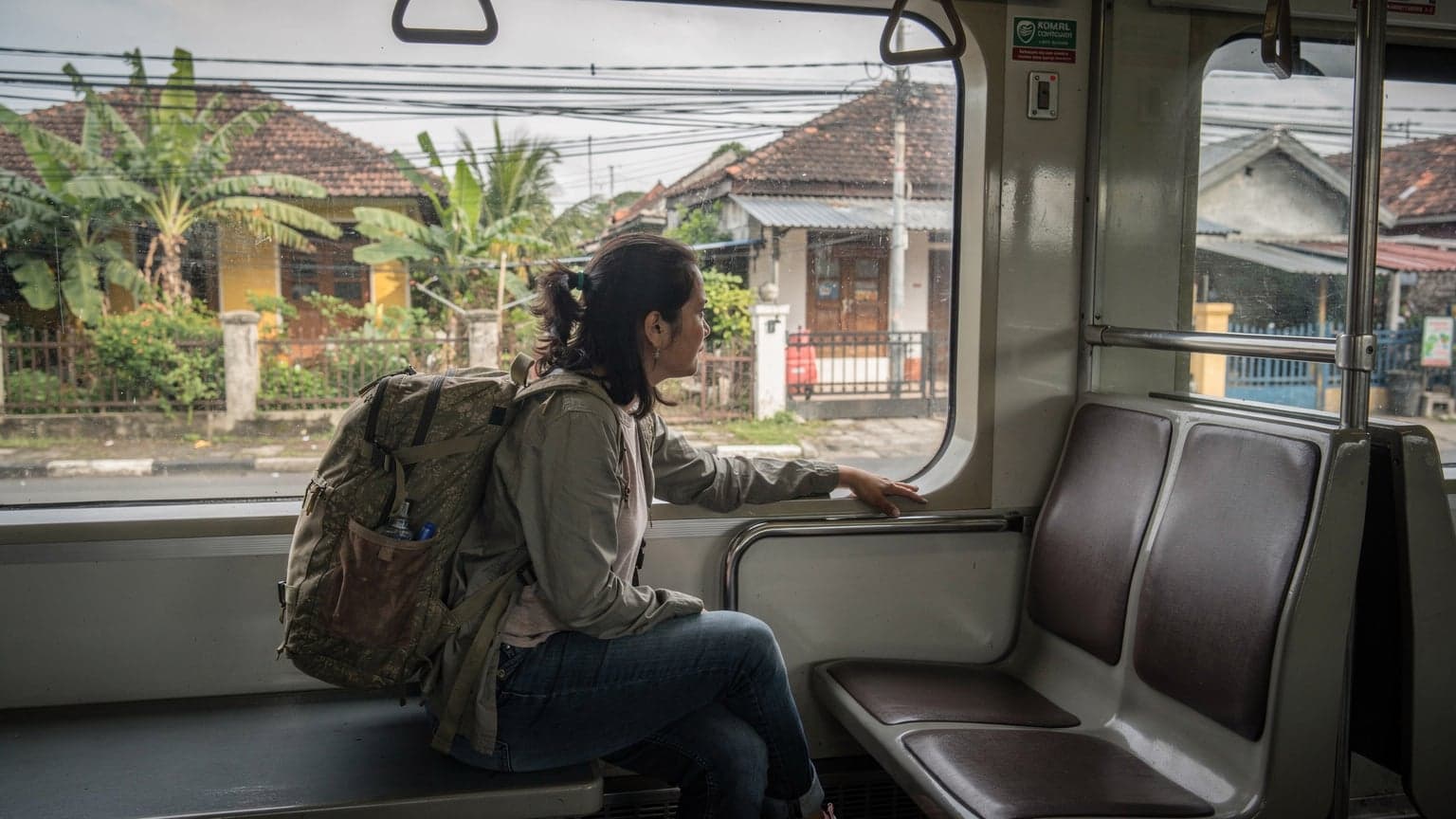 A traveler gazes thoughtfully out of a train window on the KRL Commuterline, watching the lush, suburban Indonesian landscape slide by, a backpack resting on the seat beside them.